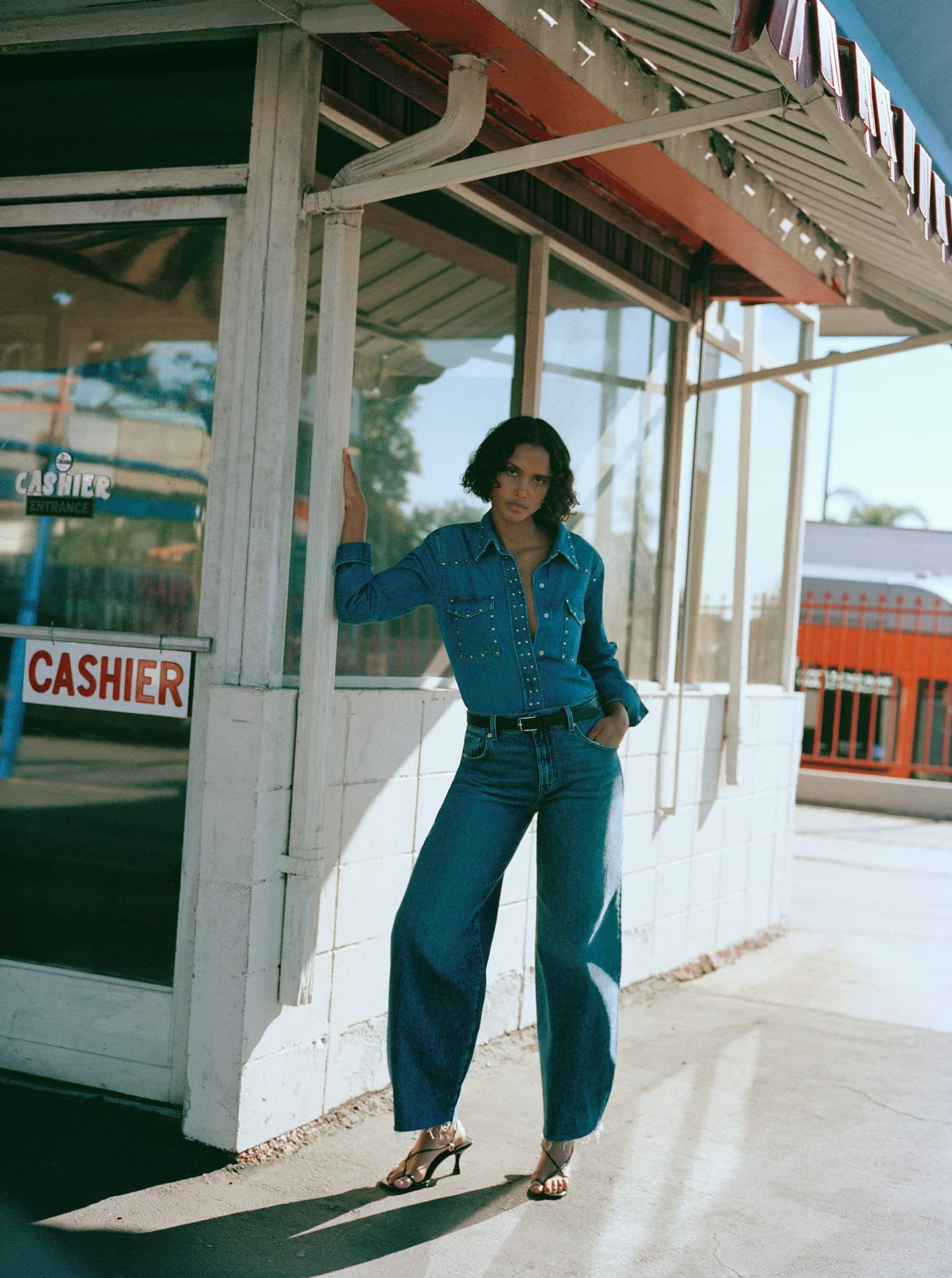 A woman poses wearing a denim shirt and jeans, standing against a vintage white building with a red 'Cashier' sign in daylight.