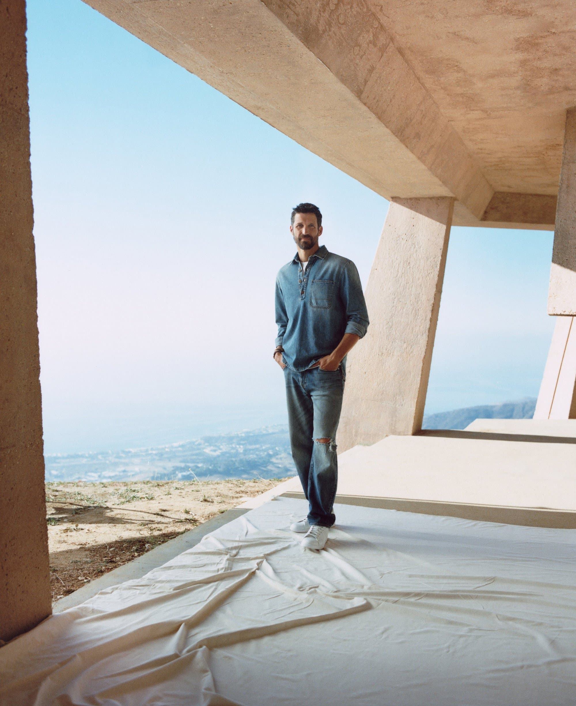 Man in denim shirt and jeans poses in a modern architectural space with an open view of a coastal landscape and bright blue sky.