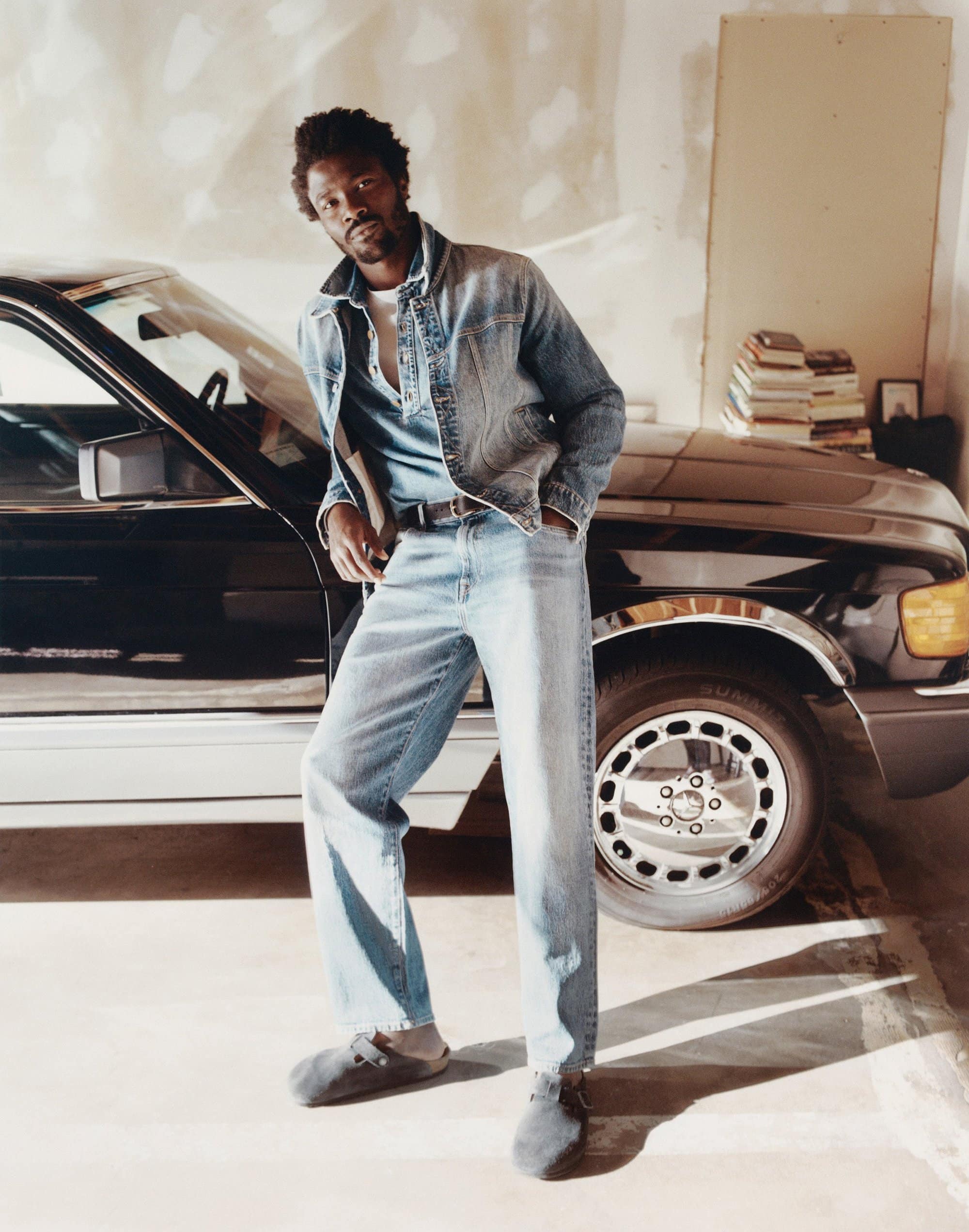 An adult male poses in a denim jacket and jeans, standing near a classic black car in an indoor setting with soft, natural lighting.
