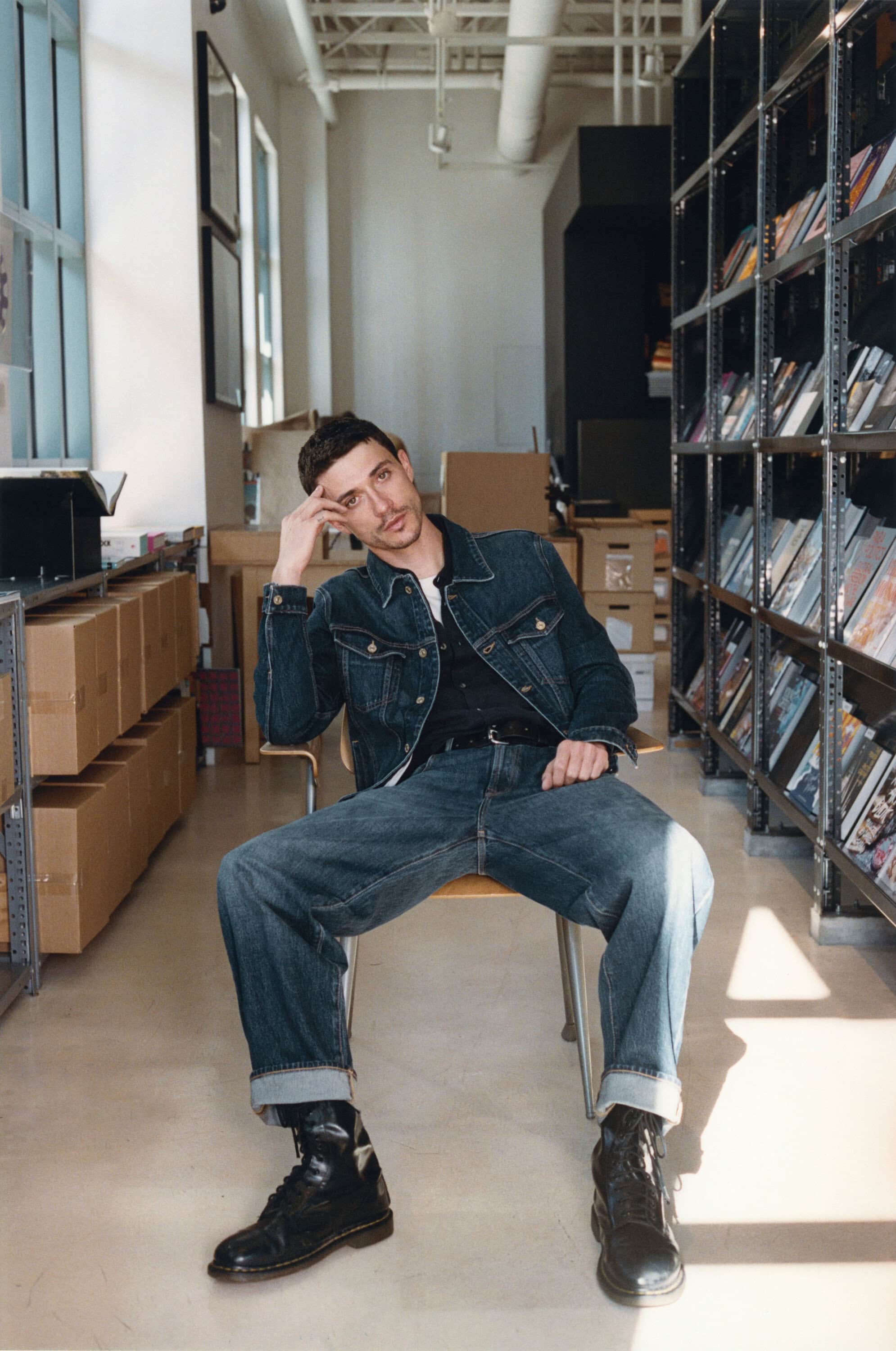 Man in denim jacket and jeans sitting on a chair in an indoor space with shelves, wearing leather boots and looking contemplative.