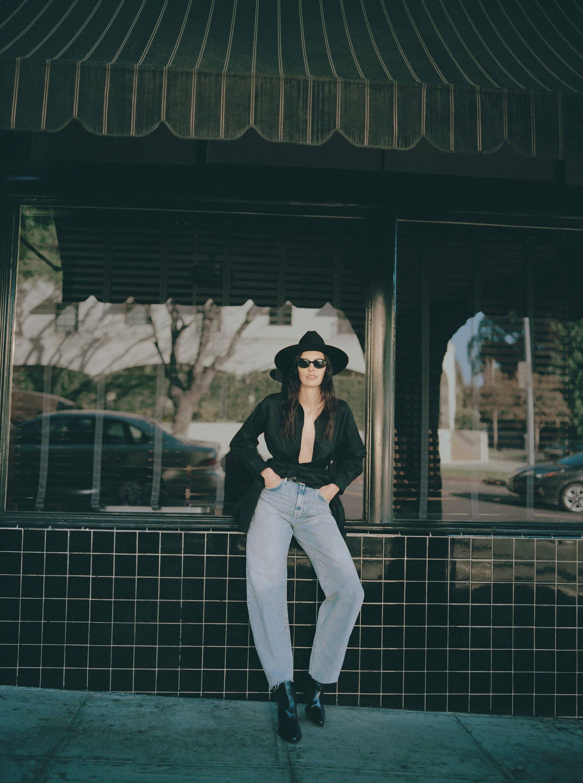 Stylish woman in hat and jeans poses against tiled building on city street, showcasing fashion and urban lifestyle in daylight.