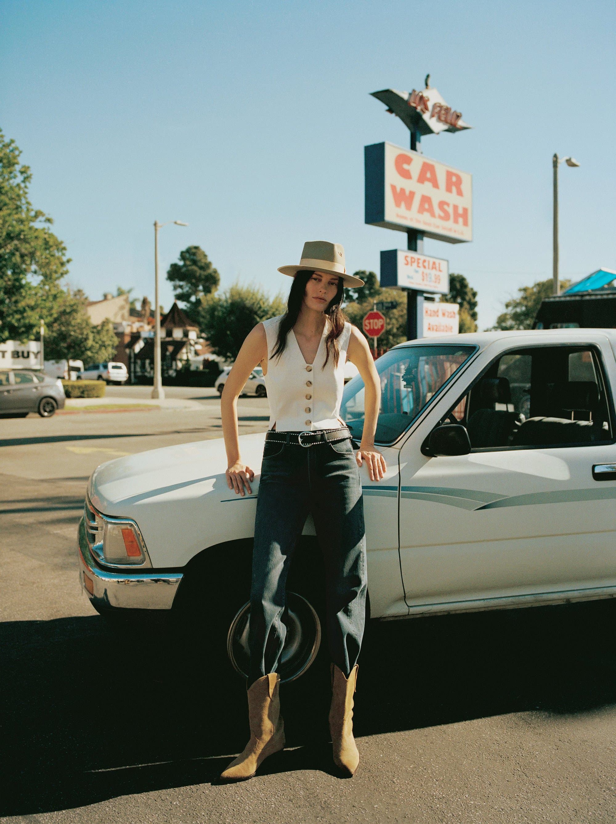 A woman poses next to a vintage pickup truck in front of a car wash under a bright blue sky, blending fashion with Americana.