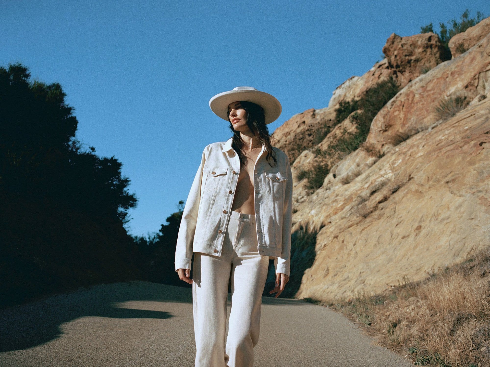 A young woman in a white denim jacket and trousers poses on a road in a desert landscape on a sunny day under a clear blue sky.
