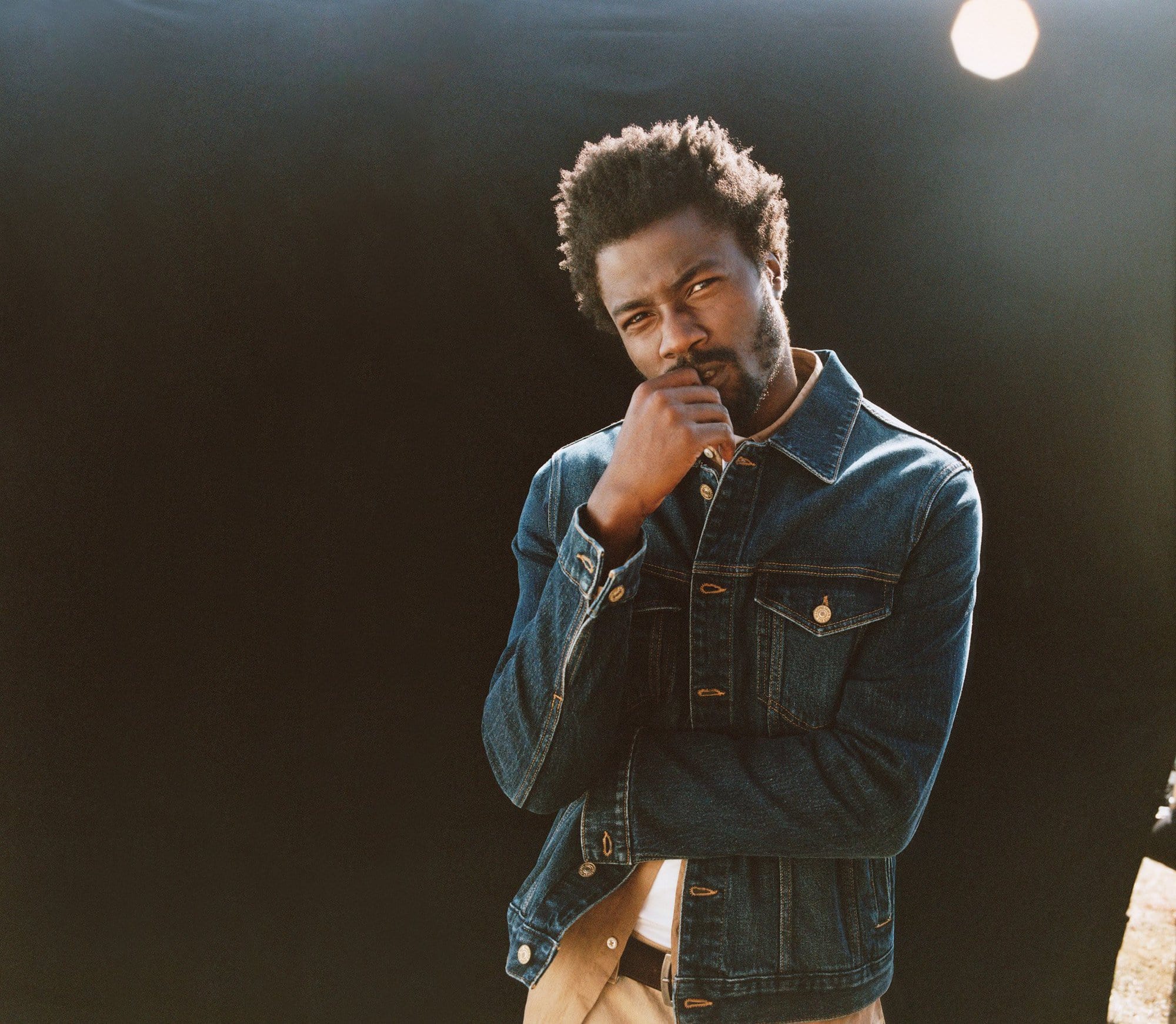 Studio portrait of an adult Black man with a beard, wearing a denim jacket, hand to chin, against a dark background with studio lighting.