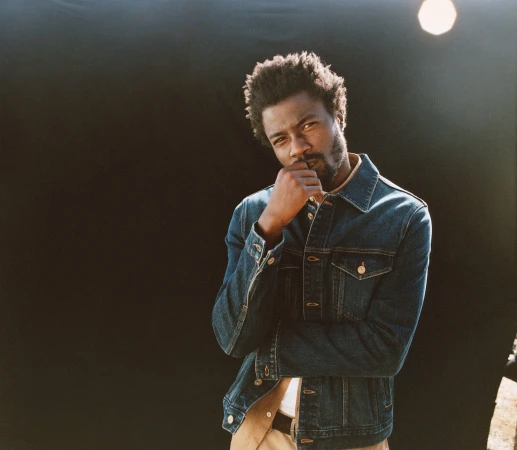 Studio portrait of an adult Black man with a beard, wearing a denim jacket, hand to chin, against a dark background with studio lighting.