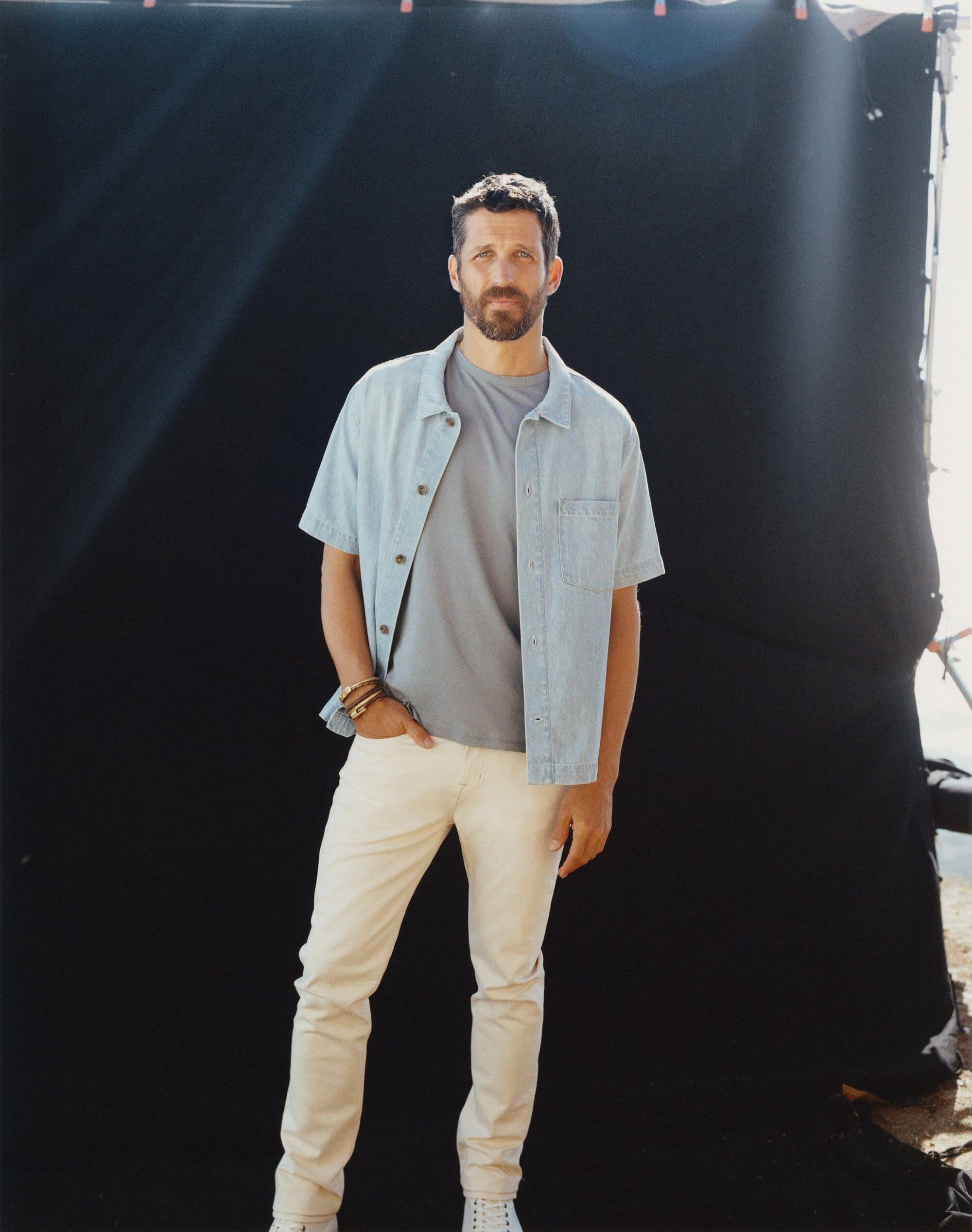 Man wearing a light blue denim shirt, grey t-shirt and light trousers standing against a black seamless backdrop.