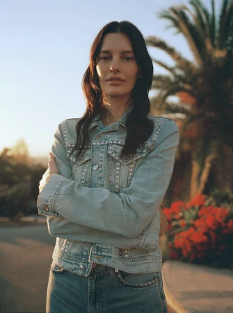 Outdoor portrait of a woman in a denim jacket and jeans with arms crossed in front of a palm tree and other foliage.