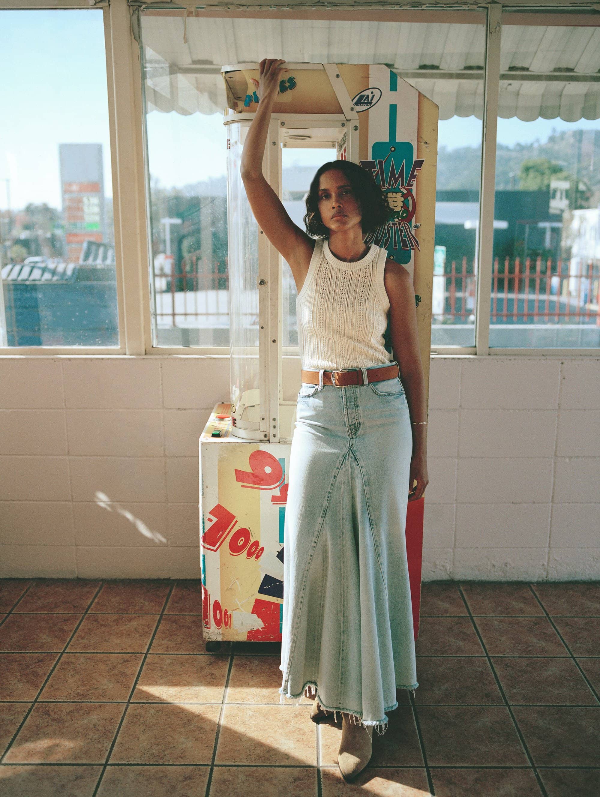 A young woman poses in front of a vintage arcade game with a denim skirt, white top, and brown belt in an outdoor setting.
