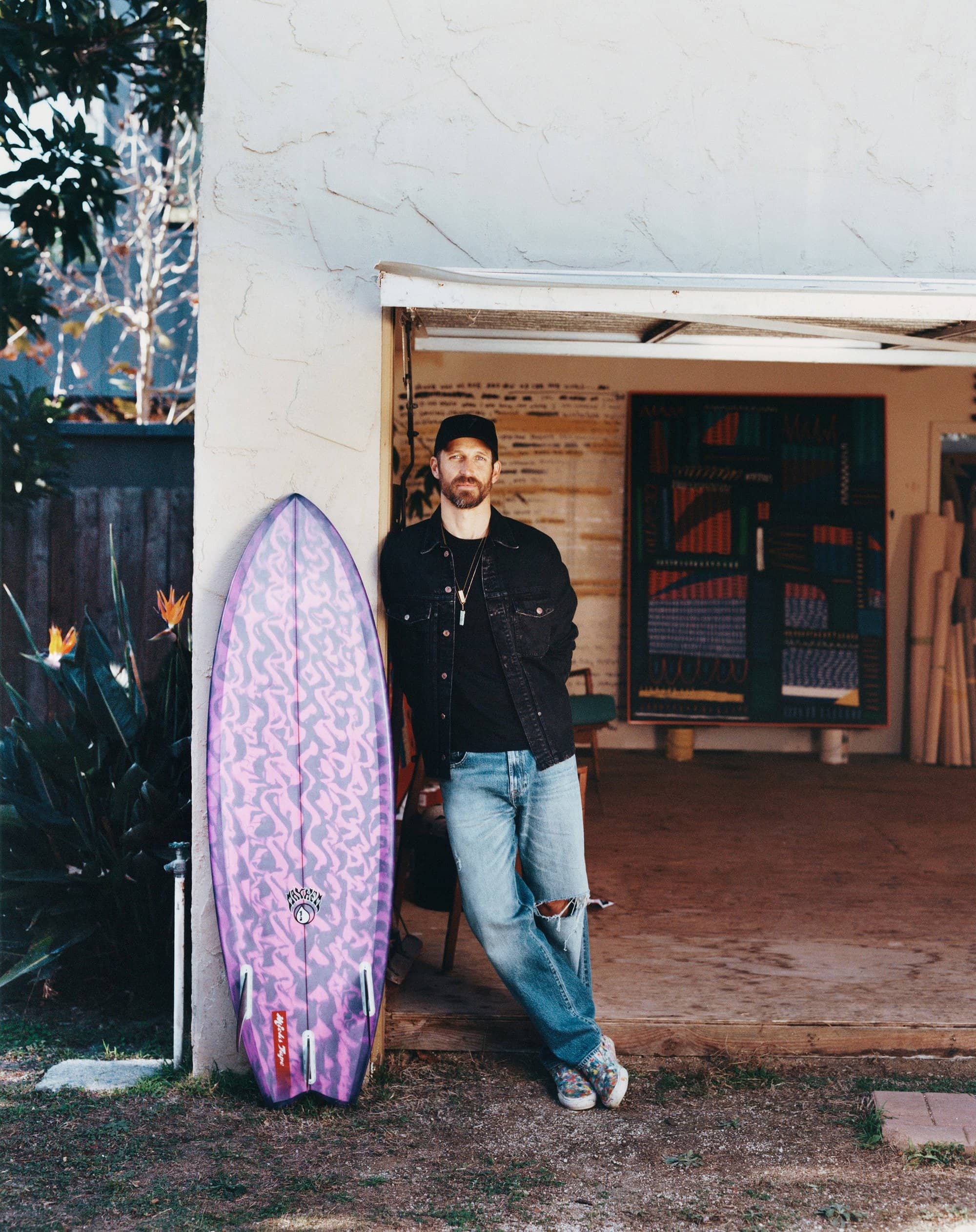 Adult male poses next to purple patterned surfboard outside a building with rolled materials and colorful artwork visible inside.