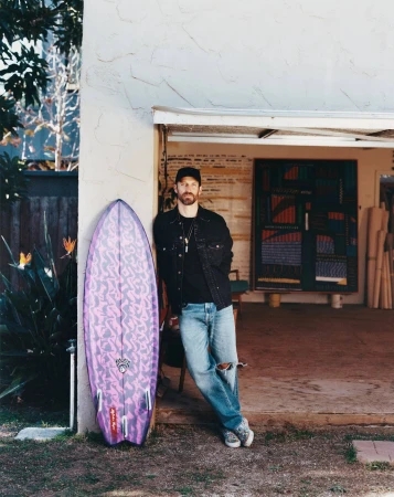 Adult male poses next to purple patterned surfboard outside a building with rolled materials and colorful artwork visible inside.