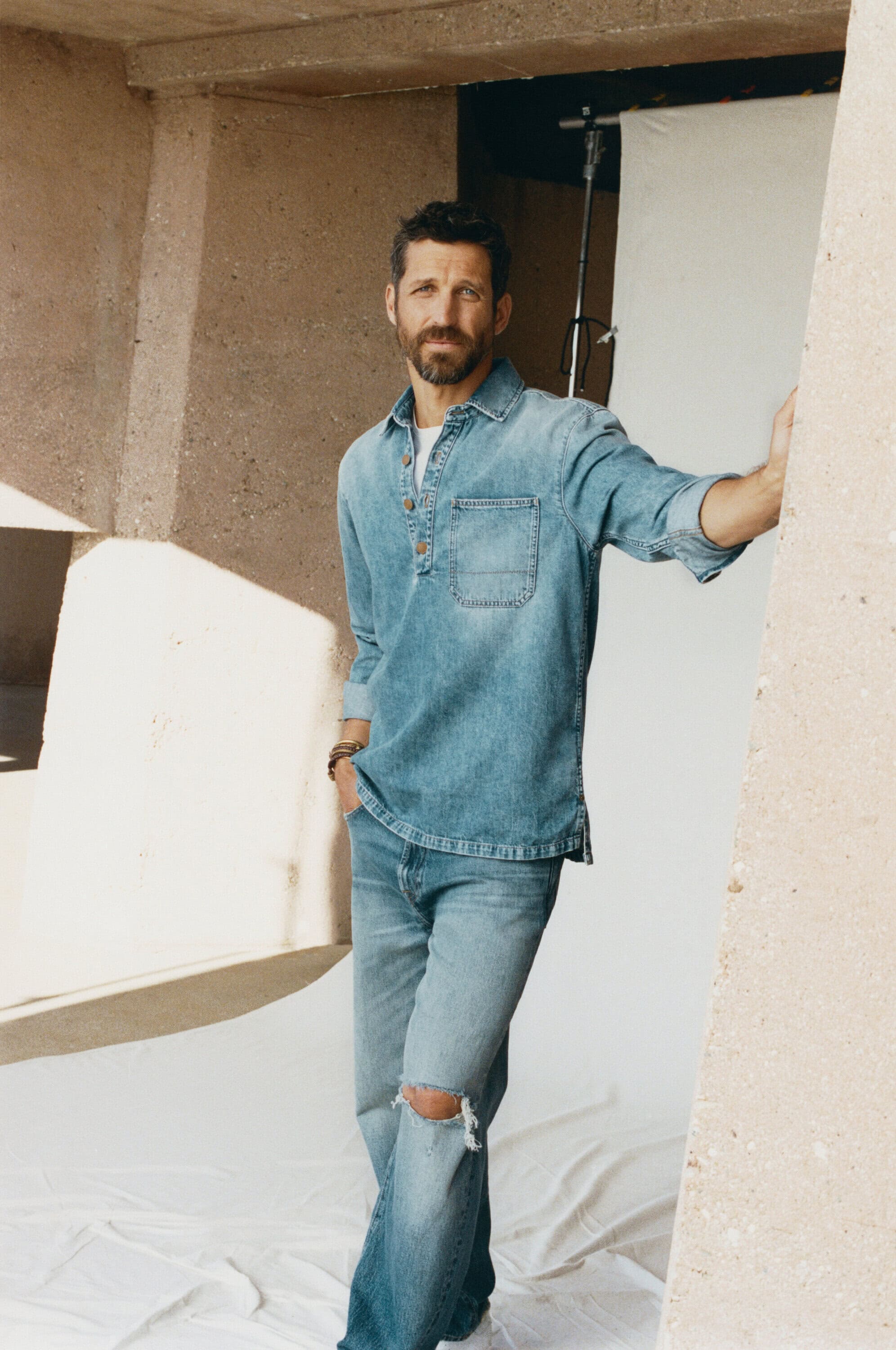 Adult male in denim shirt and jeans poses in a photo studio with white backdrop in front of a concrete wall for a fashion shot.