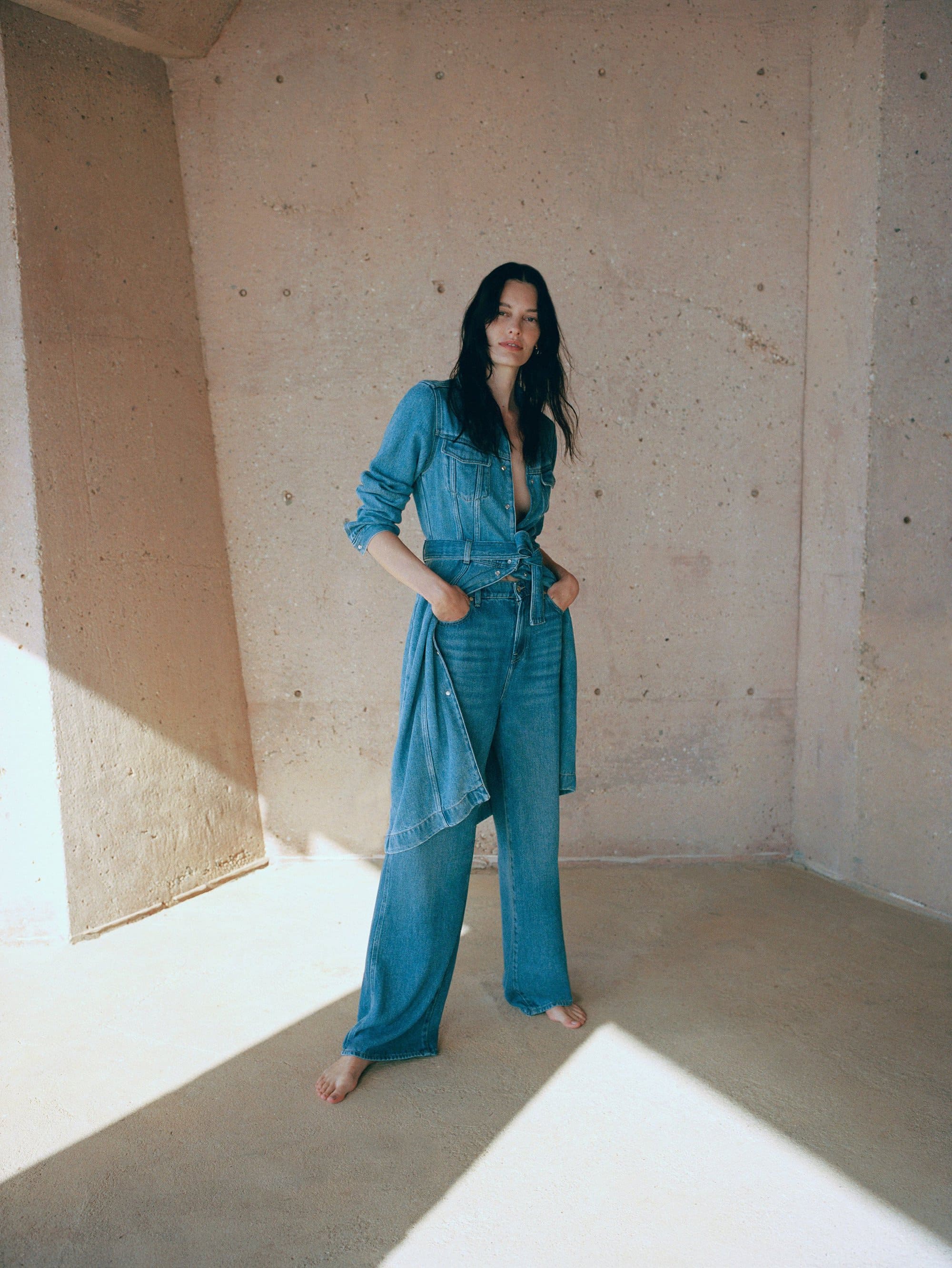 A woman in a denim shirt and wide-leg pants poses indoors against a concrete wall, highlighted by a light beam, showcasing fashion-forward simplicity.