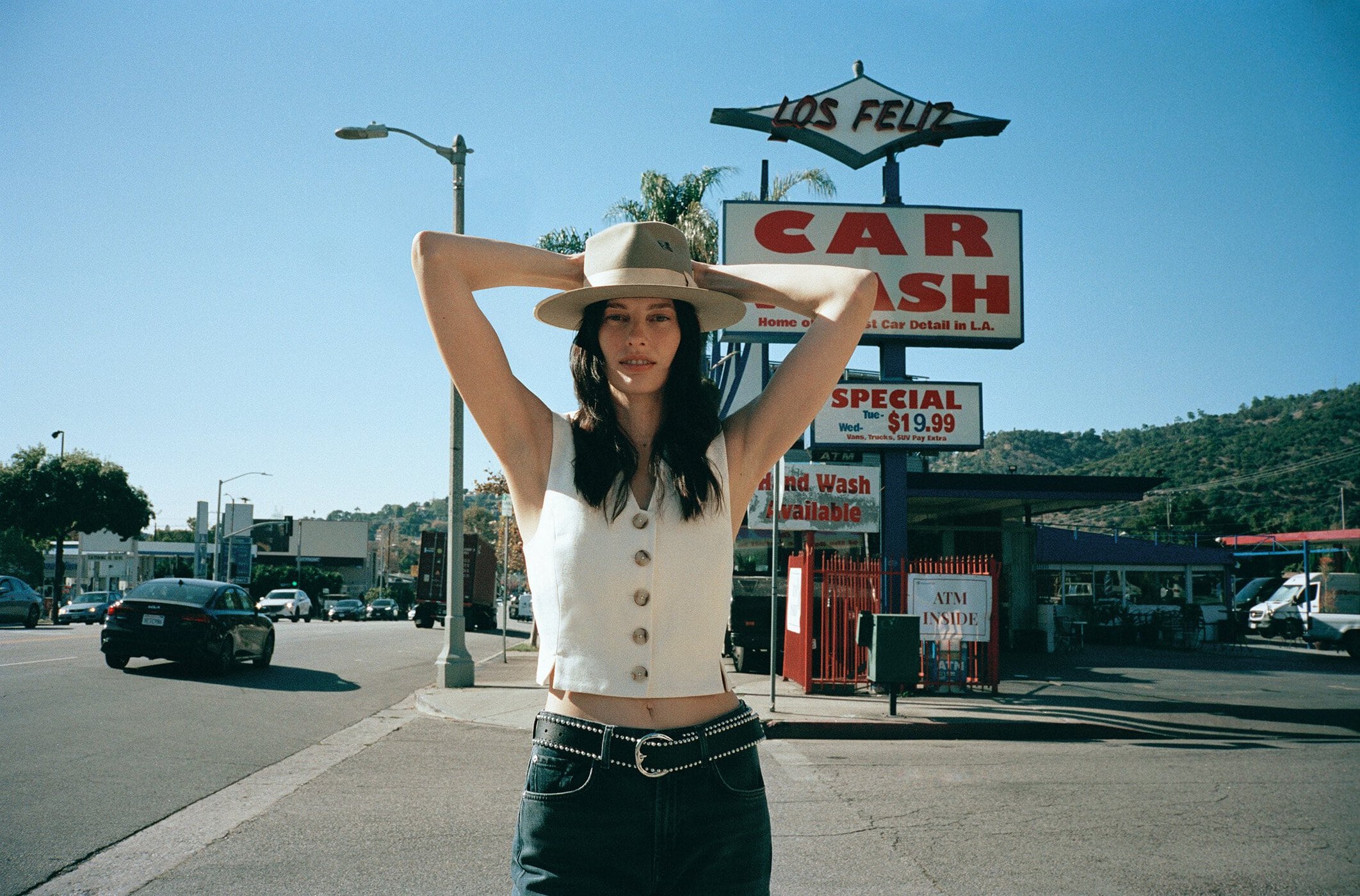 A woman in a hat and vest poses on a street corner in front of a Los Feliz car wash, with a sunlit sky and city backdrop.
