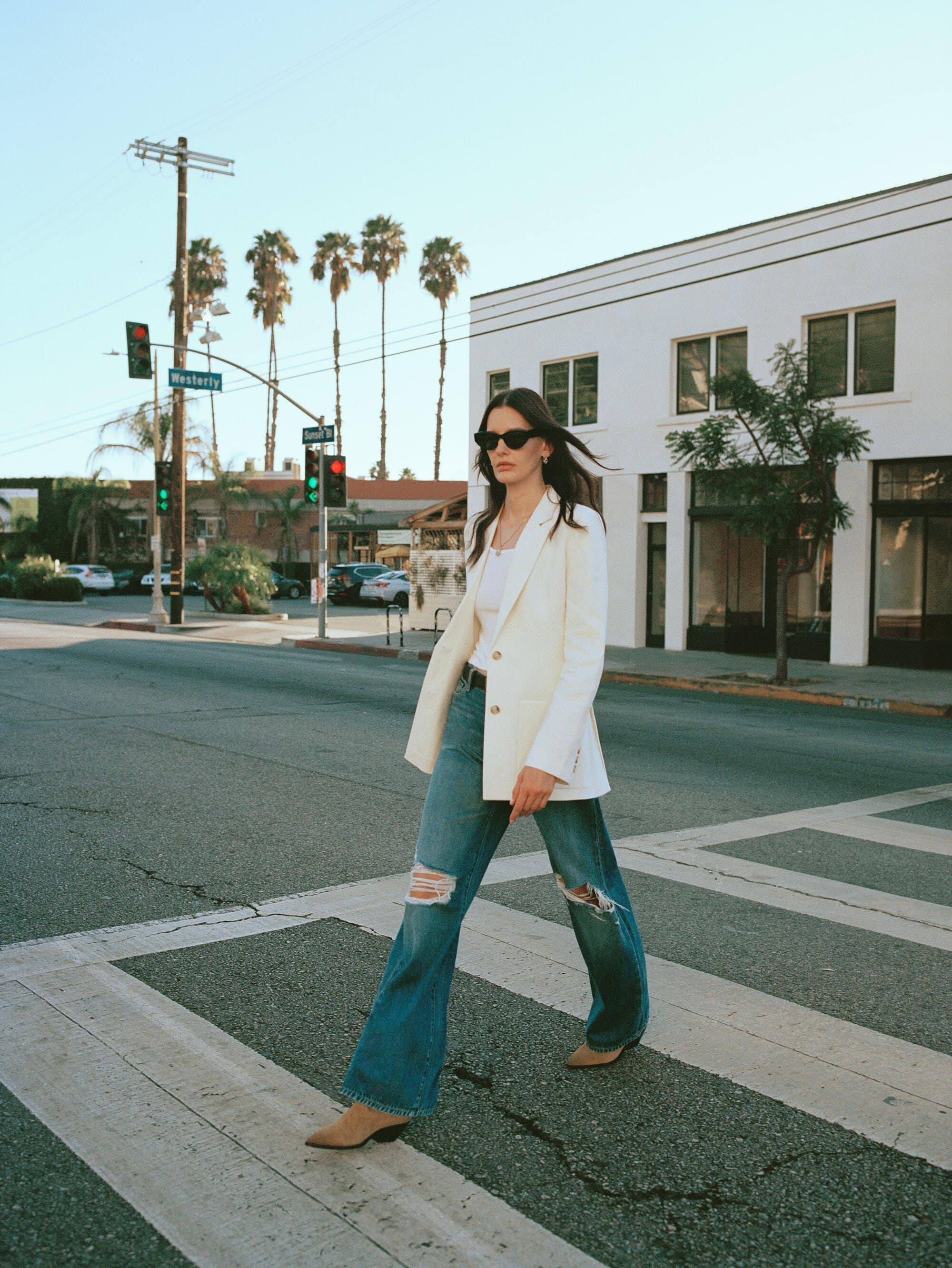 Woman in white blazer and ripped jeans crossing street on crosswalk with palm trees and buildings in the background on a sunny day.
