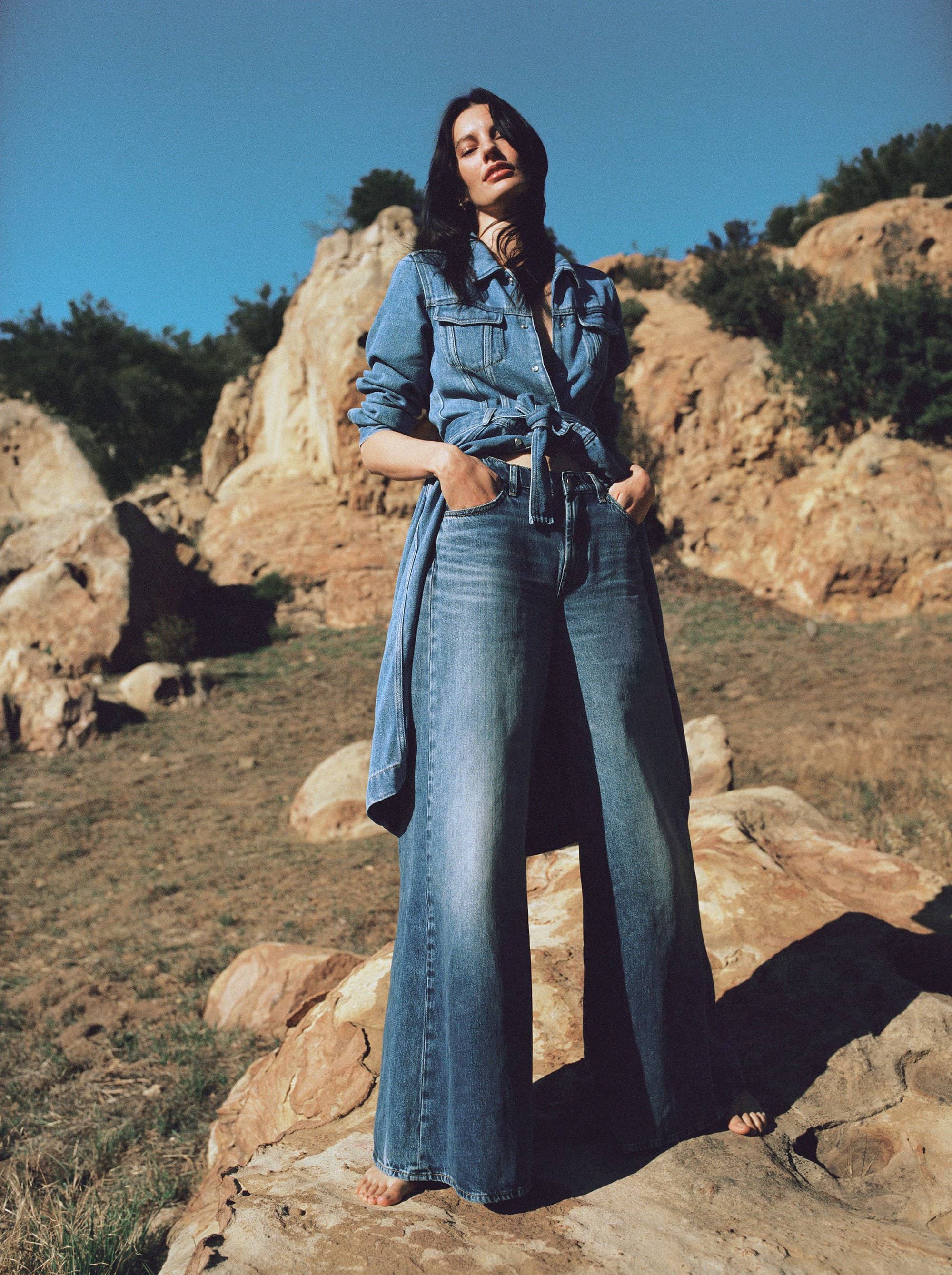 A model with long dark hair stands barefoot on a desert rock wearing a denim shirt tied at the waist and wide-leg blue jeans.