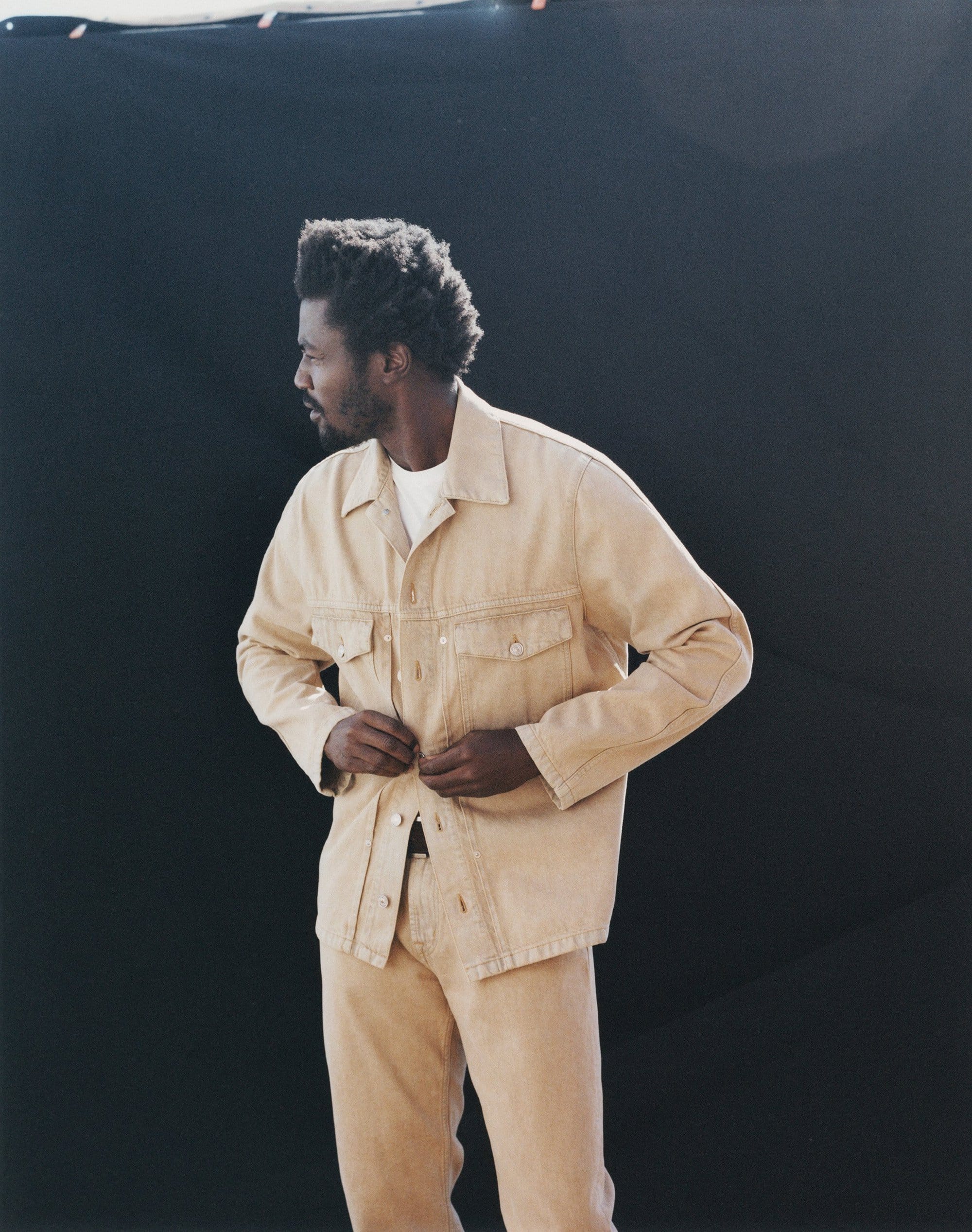Adult male in a beige denim jacket and matching pants, poses against a dark background, with a three-quarter view looking left.