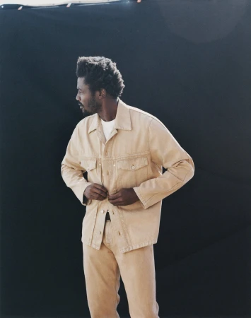 Adult male in a beige denim jacket and matching pants, poses against a dark background, with a three-quarter view looking left.