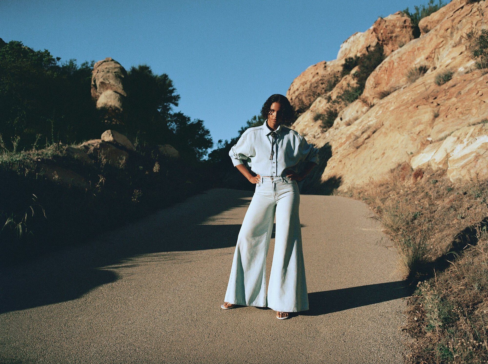 Woman in denim outfit posing on path between rocky hills under blue sky; outdoor fashion editorial on a sunny day.