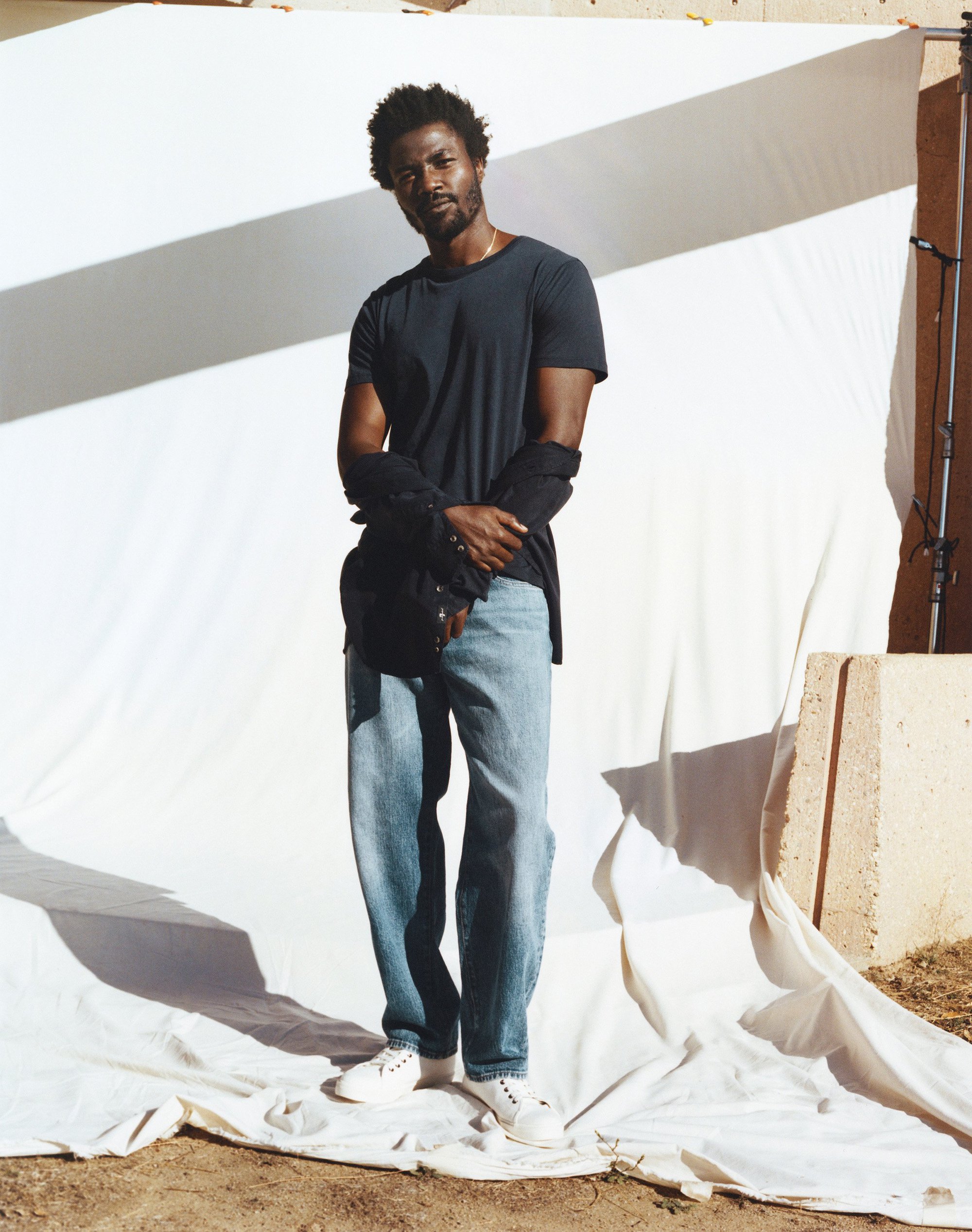 Man in casual wear stands against a white backdrop, wearing jeans, a dark t-shirt, and a jacket tied around his waist in a studio setting.