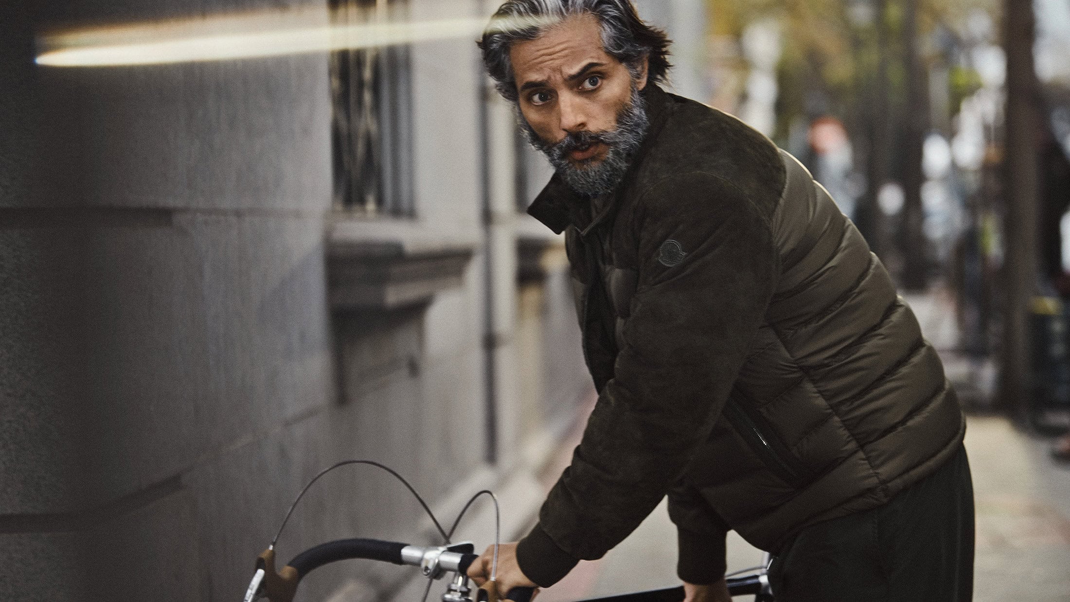 A man with gray hair and a beard poses outside on a city street wearing a puffer jacket and holding a bicycle handlebar.