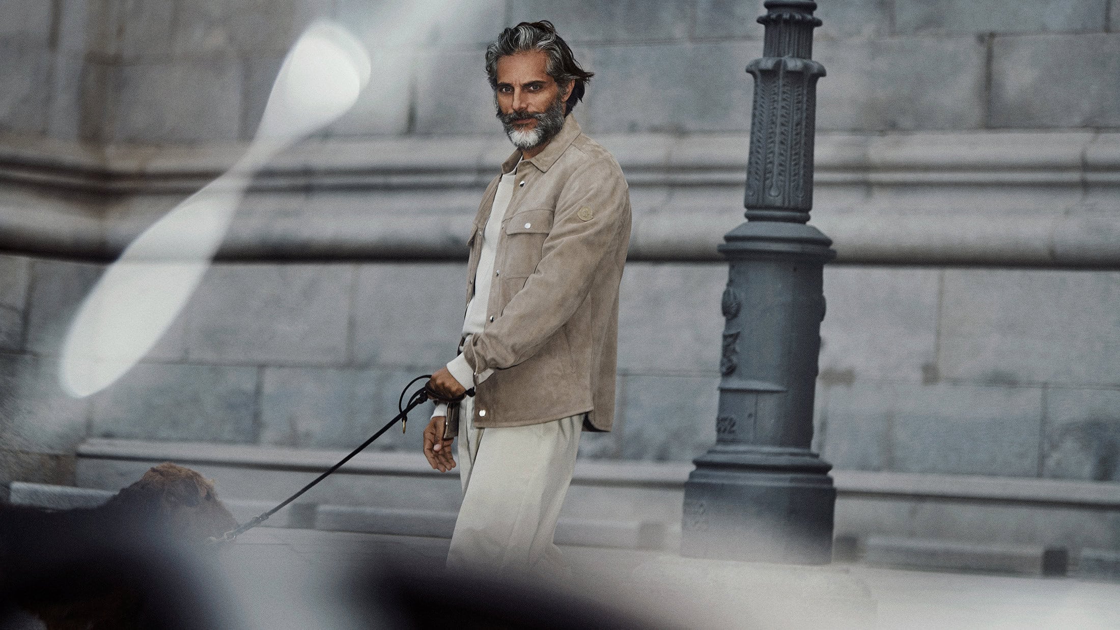 Mature man with grey hair and beard walks his dog along a city sidewalk near a brick wall and black metal light post.