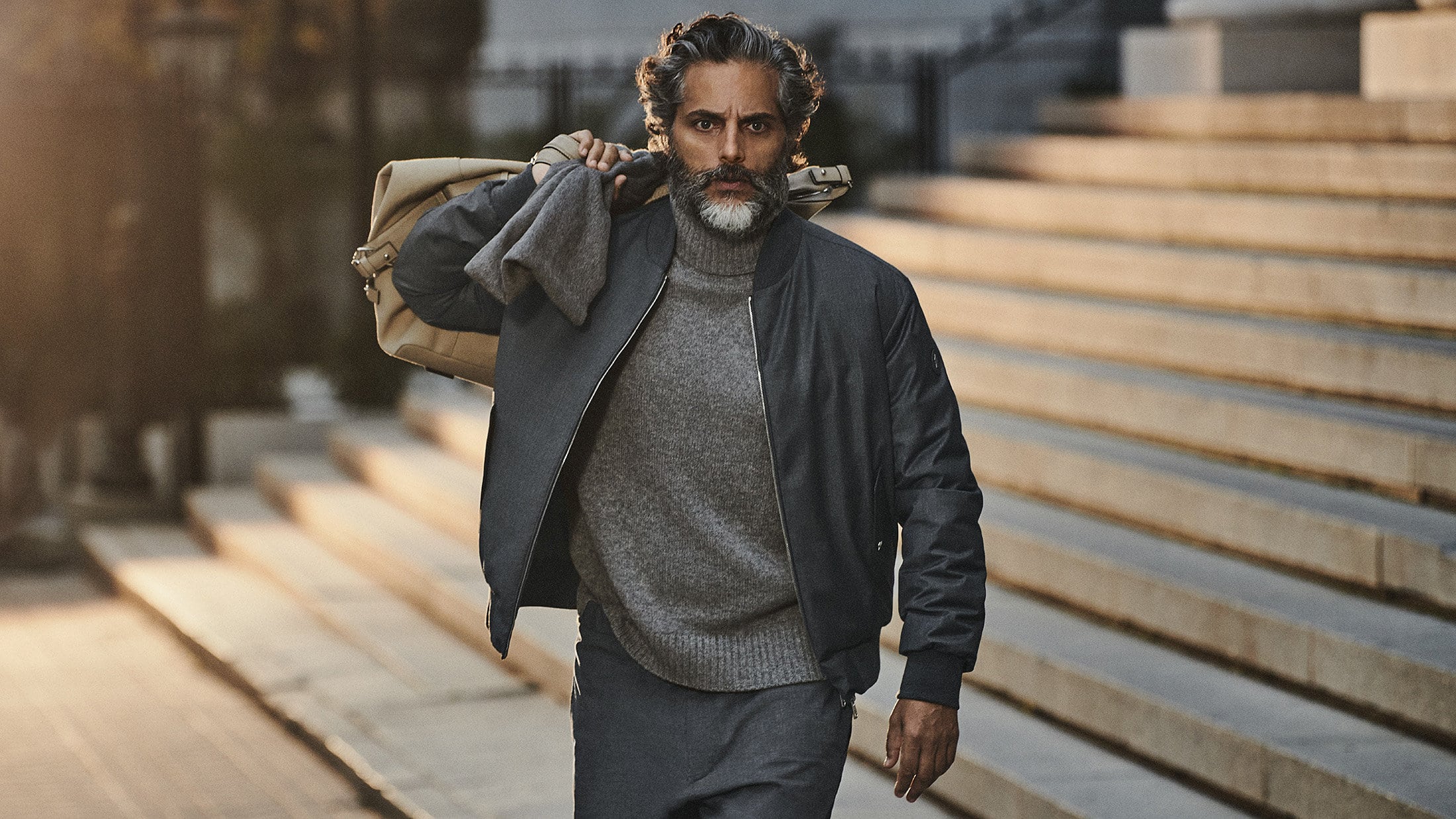 A middle-aged man with a beard walks down stone steps carrying a duffel bag. He is wearing a gray sweater and a dark bomber jacket.