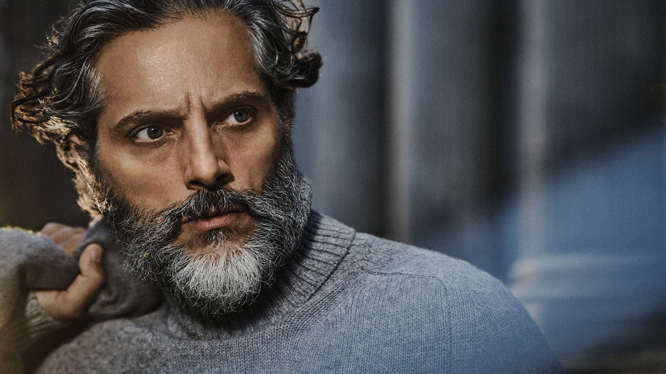 Close-up portrait of a mature man with graying hair and a beard, wearing a gray turtleneck sweater, looking off to the side against a neutral backdrop.