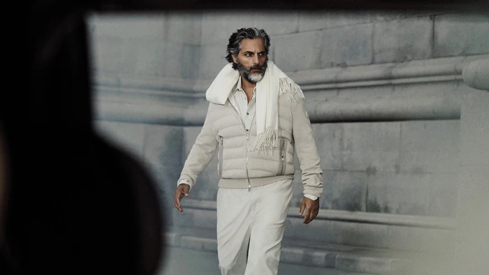 Joaquín Furriel in a white puffer jacket and scarf walking outdoors in front of a stone building facade