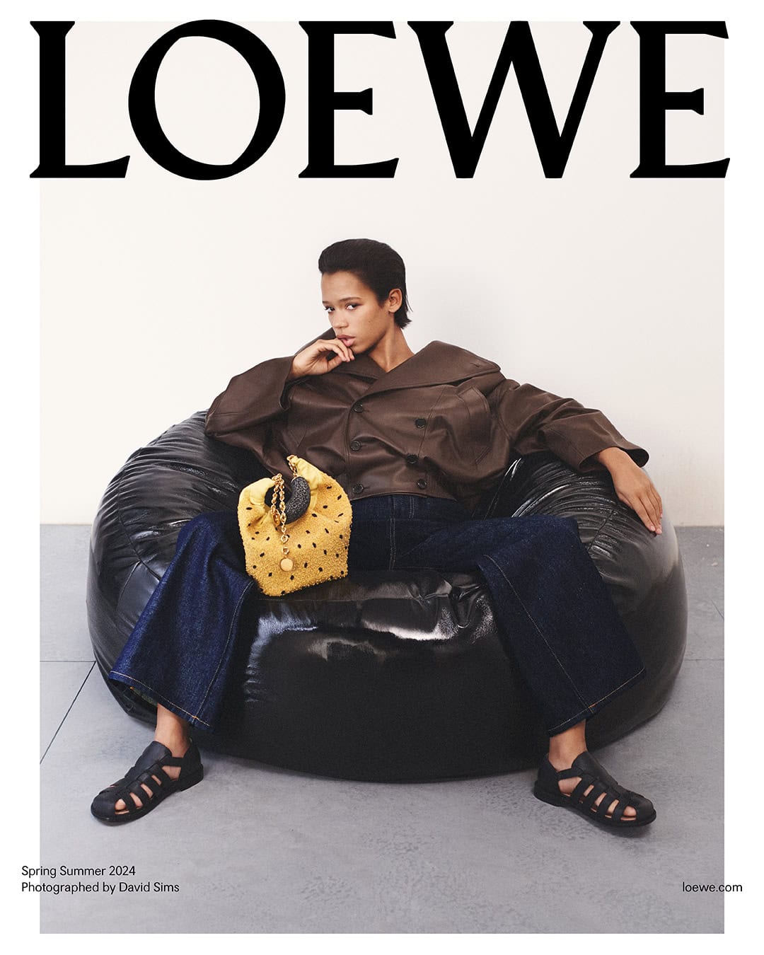 A fashion editorial-style photograph presents a female model in a brown jacket and blue jeans, seated on a black beanbag chair, holding a purse.
