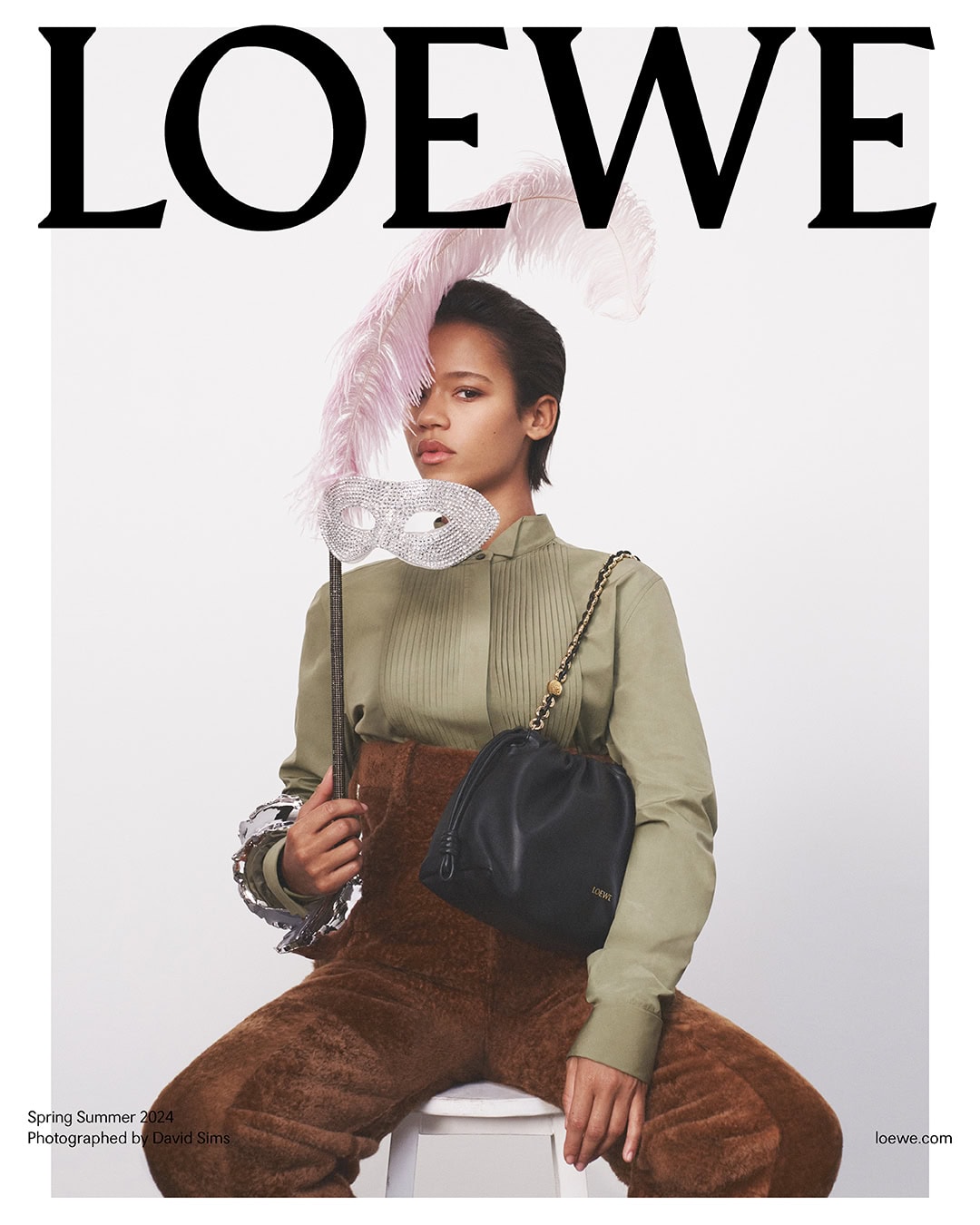Fashion editorial shot featuring a person wearing a green top and brown pants, accessorized with a black bucket bag and decorative mask.