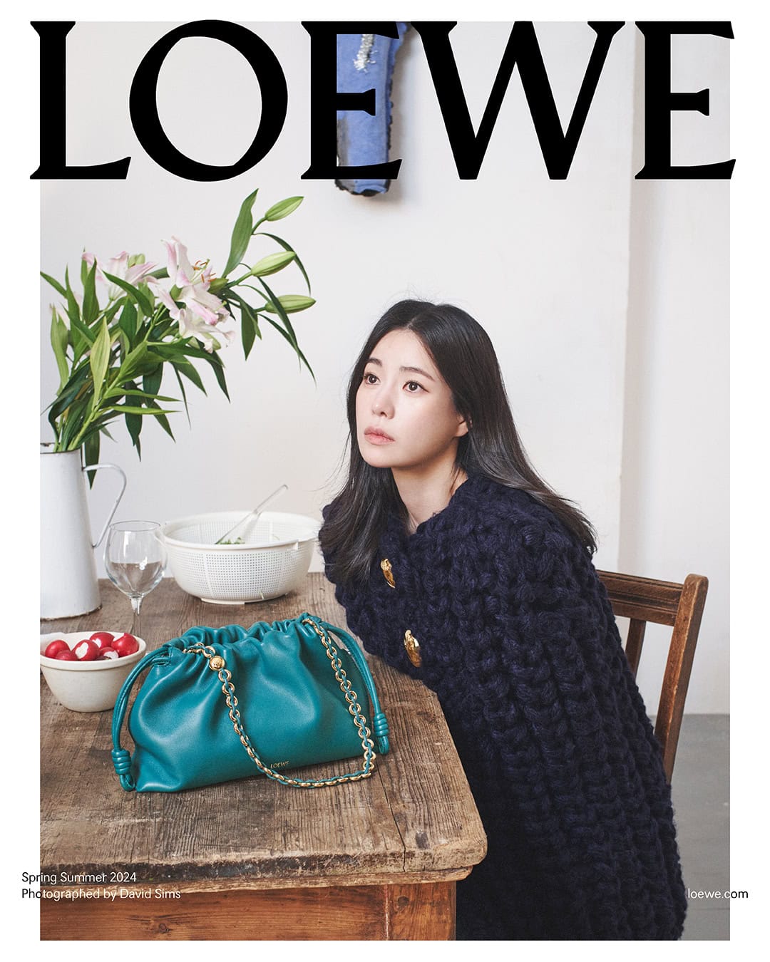 Fashion editorial featuring an Asian woman wearing a blue knit cardigan, posing next to a teal leather handbag and a flower arrangement indoors.