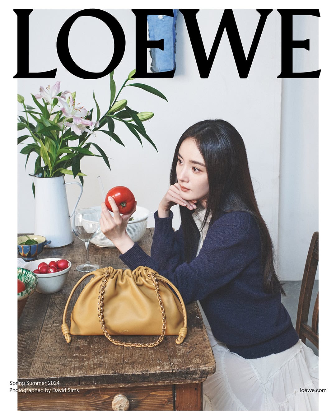 A woman sits at a table displaying a Loewe handbag, holding an apple, with flowers in a vase and the Loewe logo above her.