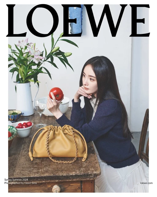A woman sits at a table displaying a Loewe handbag, holding an apple, with flowers in a vase and the Loewe logo above her.