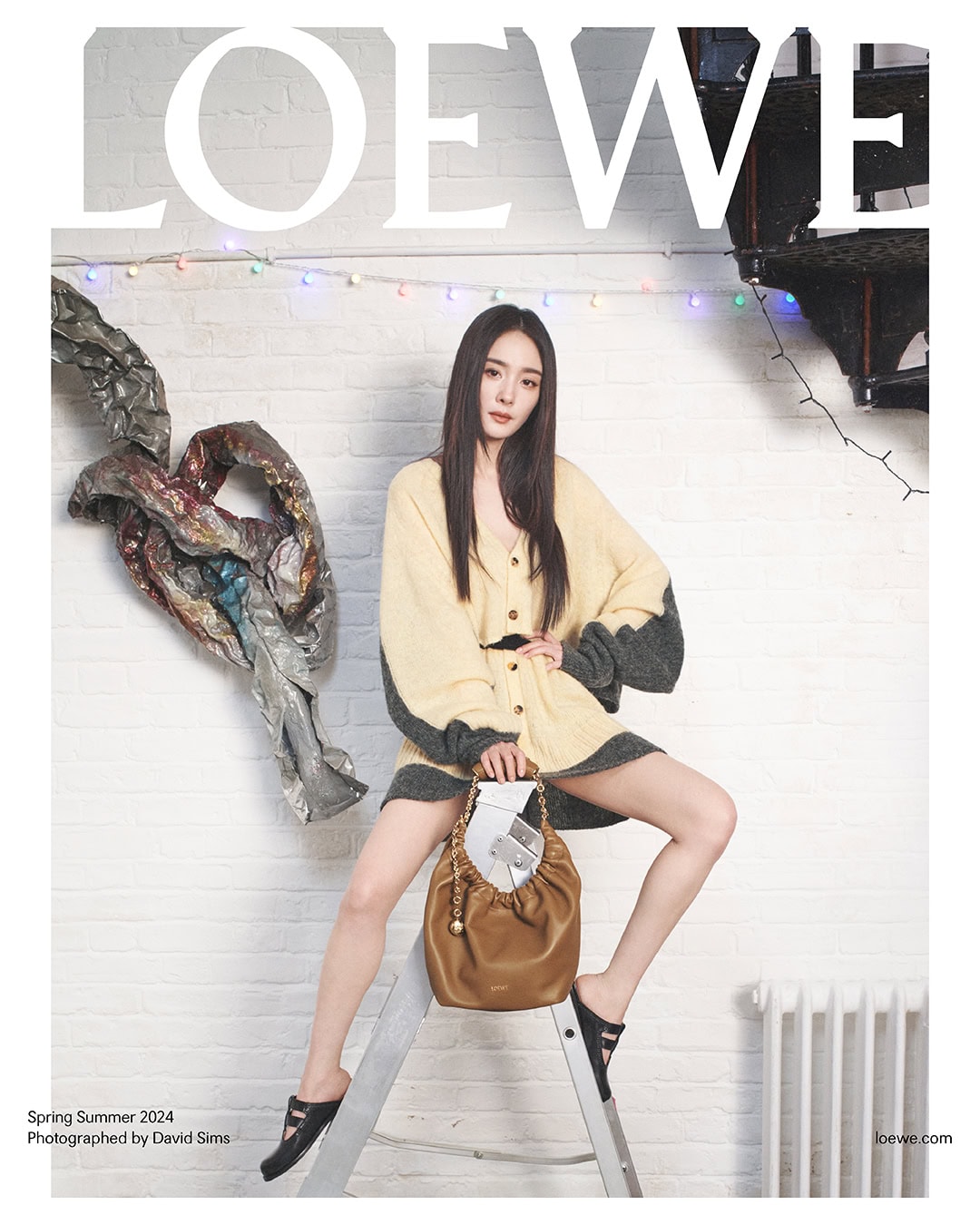 Fashion editorial featuring an adult woman in a two-tone cardigan, holding a brown purse, and posing on a silver ladder indoors.