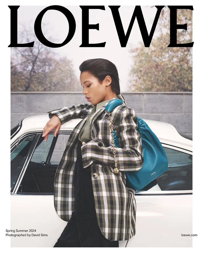 Model leaning against a white vintage car wearing a plaid blazer and carrying a teal leather handbag for Loewe