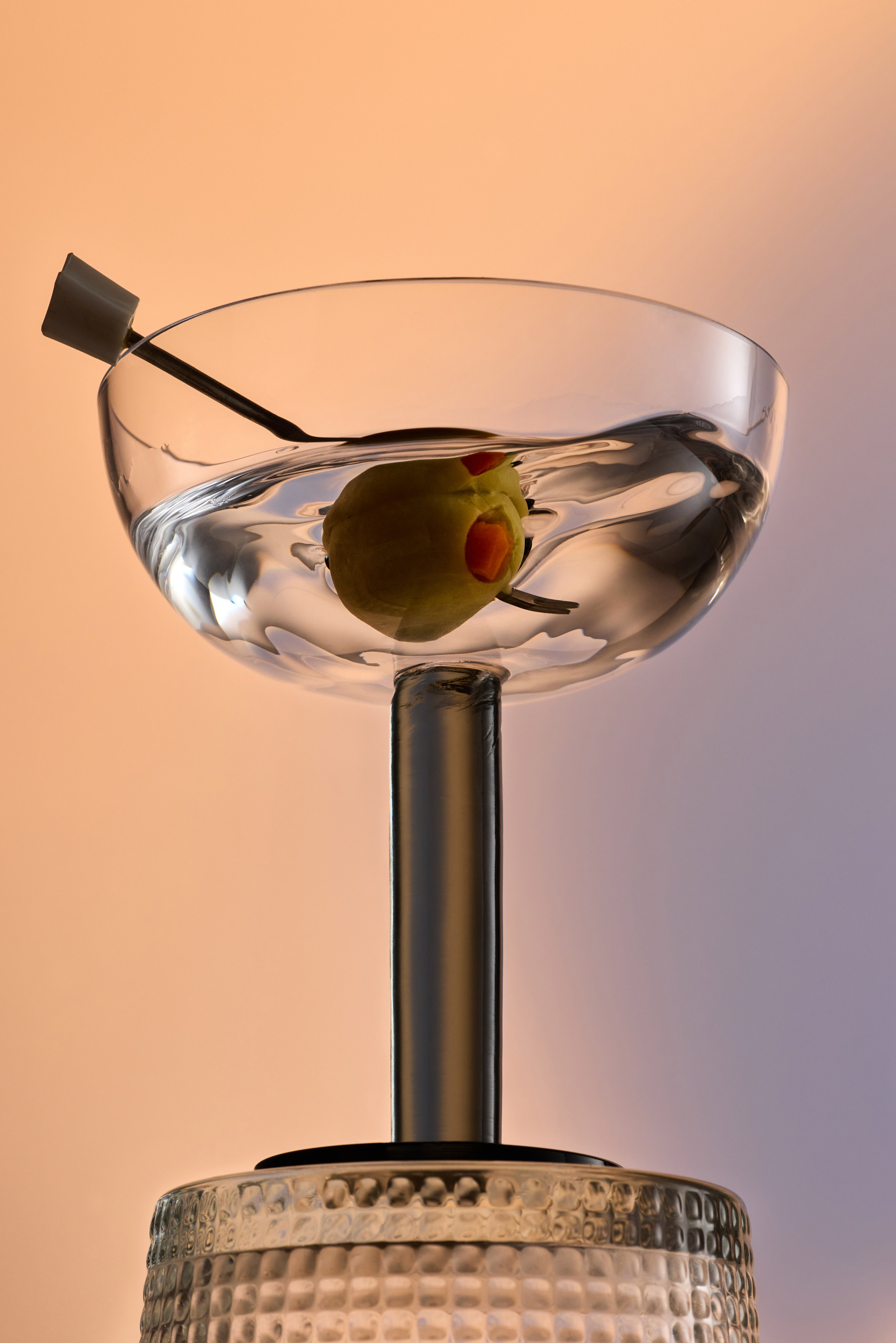 Close-up of a clear martini glass filled with liquid, containing a green olive with a pimento, illuminated by soft lighting.