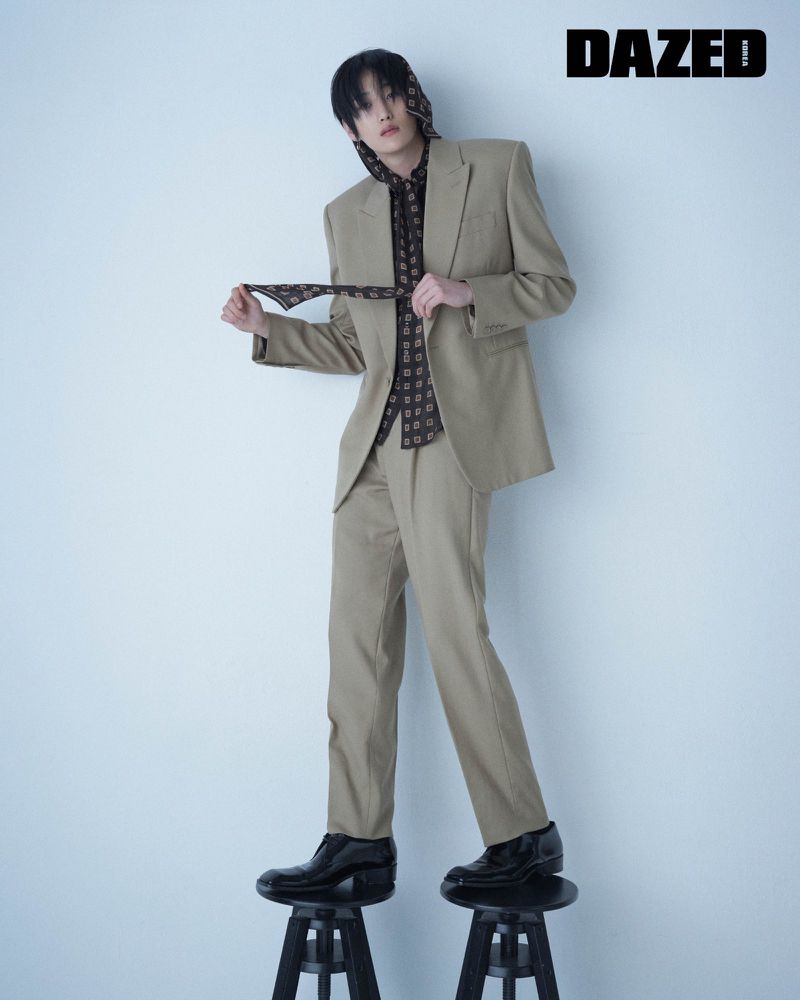 Fashion editorial shot of a slender person in a beige suit and printed headscarf standing on black stools in front of a white background.
