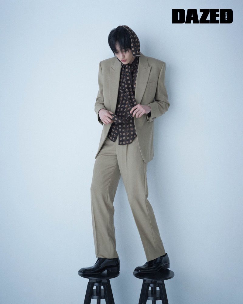 Full shot of a young adult in a tan suit and patterned shirt, standing on two black stools against a gradient blue background.
