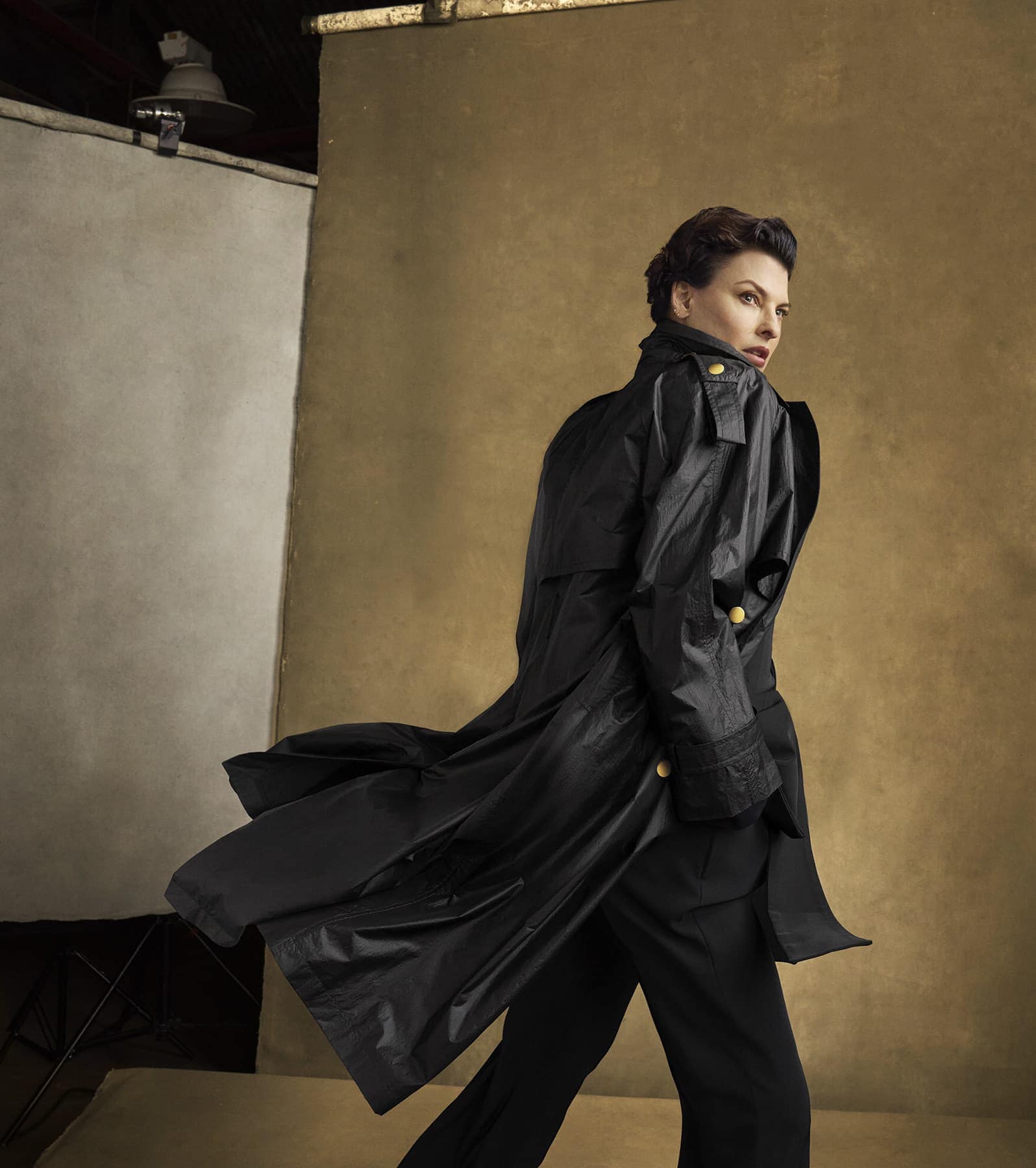 Elegant woman in a black trench coat posing in a studio against a neutral backdrop, showcasing fashion and style in a controlled environment.