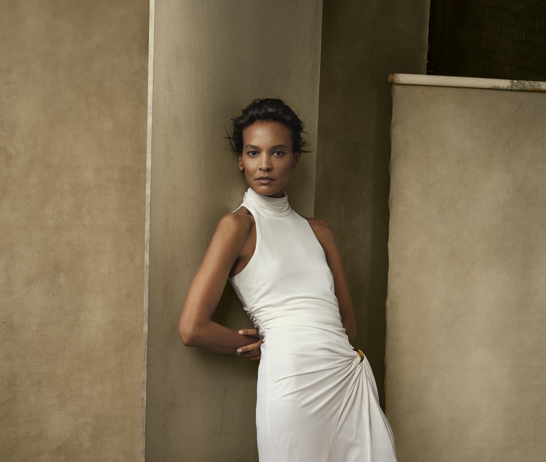 Model Liya Kebede leaning against a column wearing a white sleeveless dress for the Donna Karan Spring 2024 ad campaign.