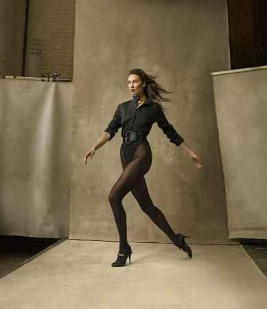 A slender woman in a black bodysuit, shirt, tights, and heels walks with arms outstretched against a neutral studio backdrop.