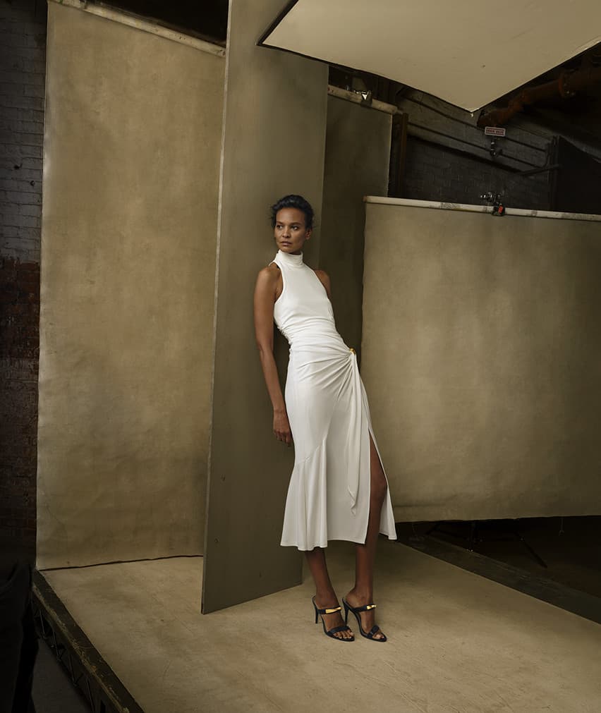 An adult female fashion model in a white dress leans against a studio backdrop. She is wearing high heels and looking left.