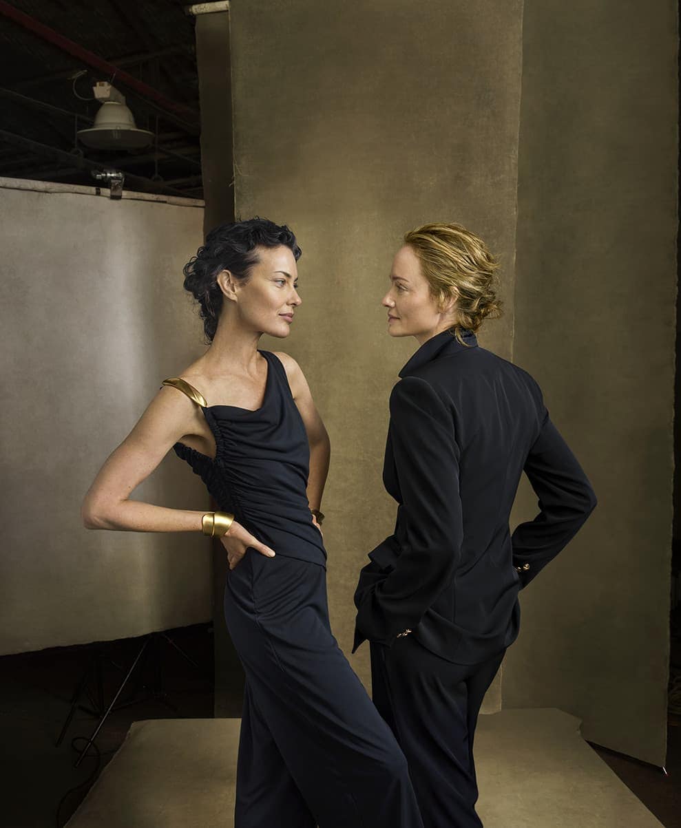 Two adult women in a photo studio wearing formal attire and jewelry. One woman is in a black dress and the other in a black suit.