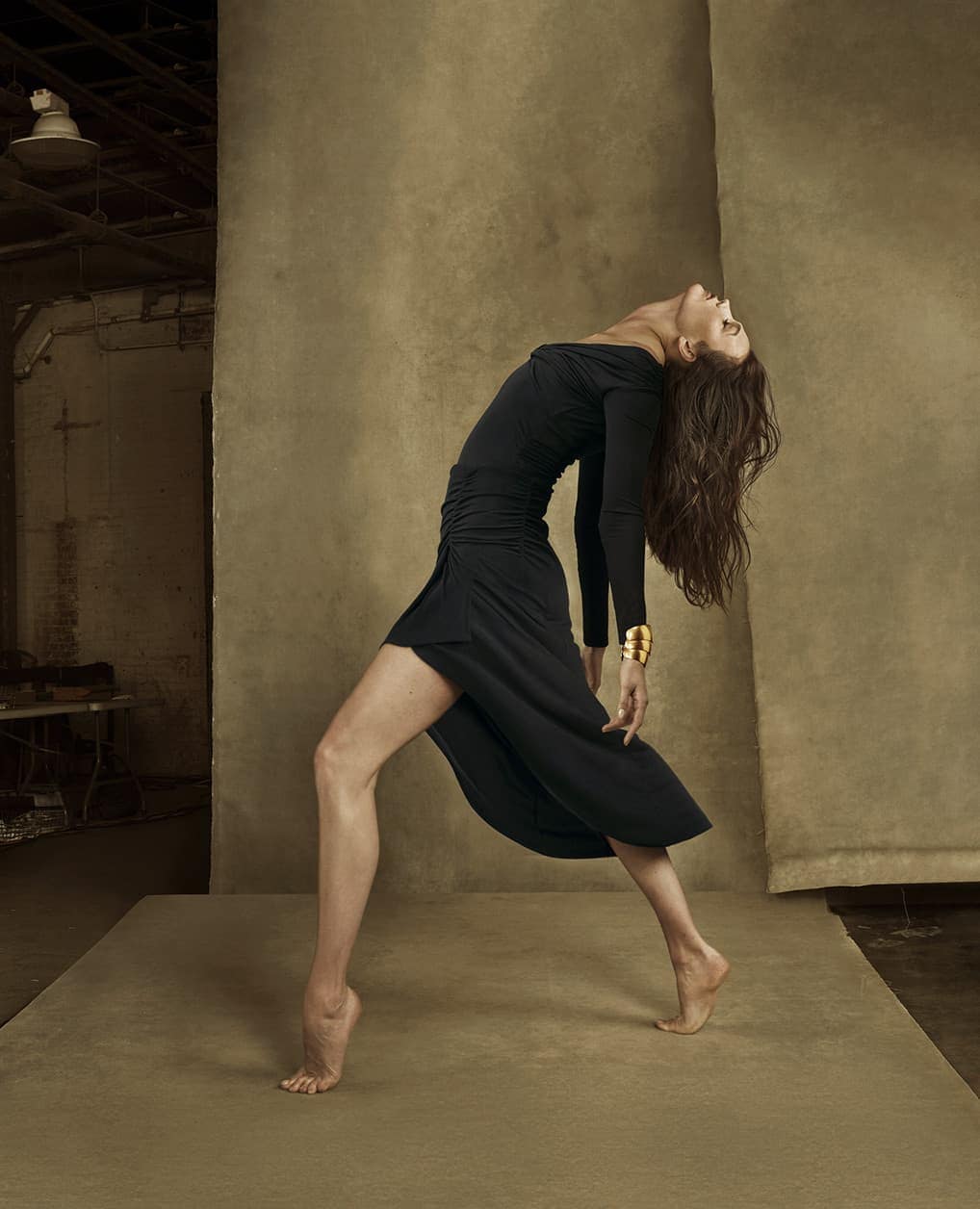 Barefoot woman in a black dress poses in a studio with textured backdrop. Her head is tilted back and her arms are outstretched.
