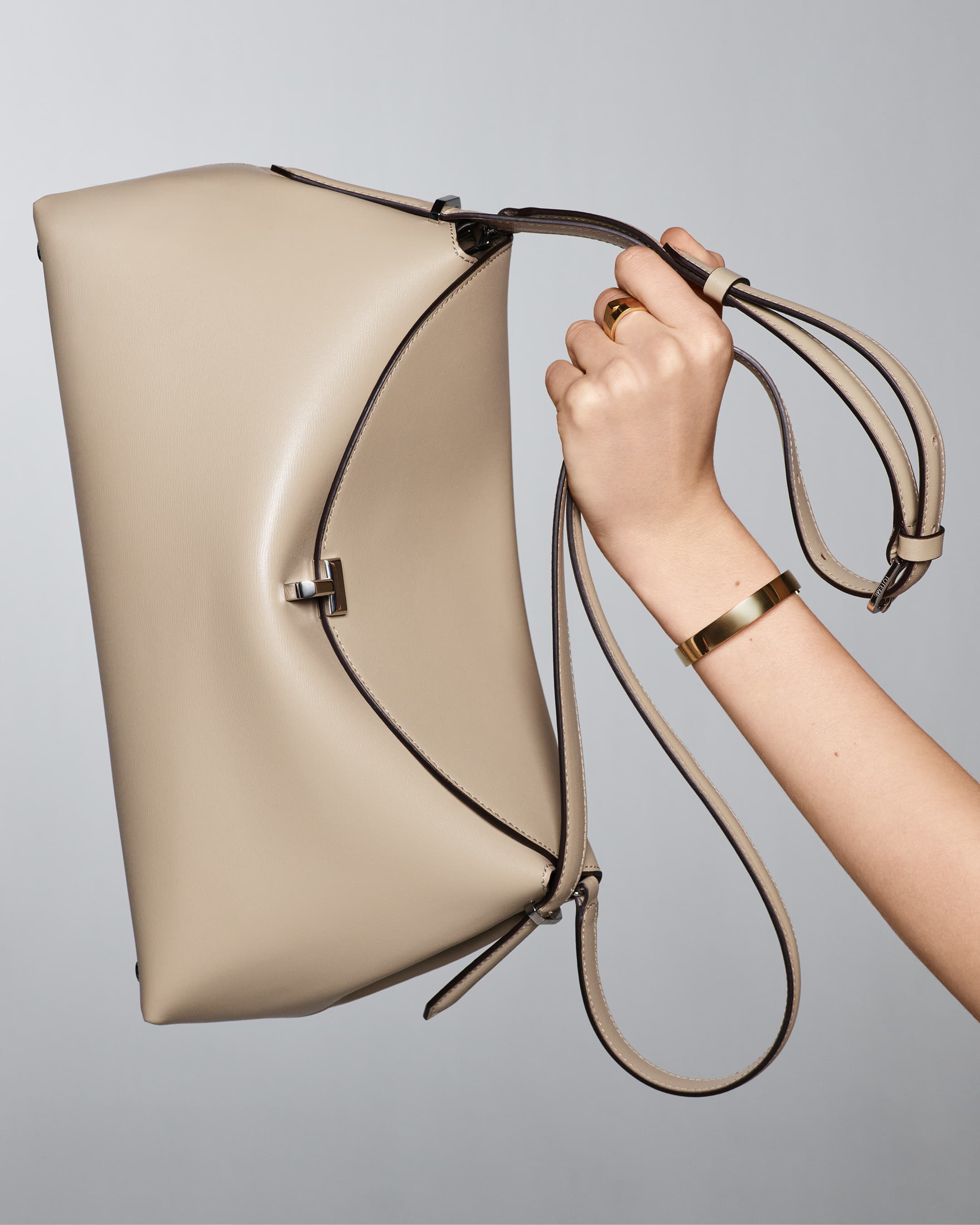 Close-up of a light beige leather handbag with a shoulder strap held by a fair-skinned adult female hand wearing a gold bangle.