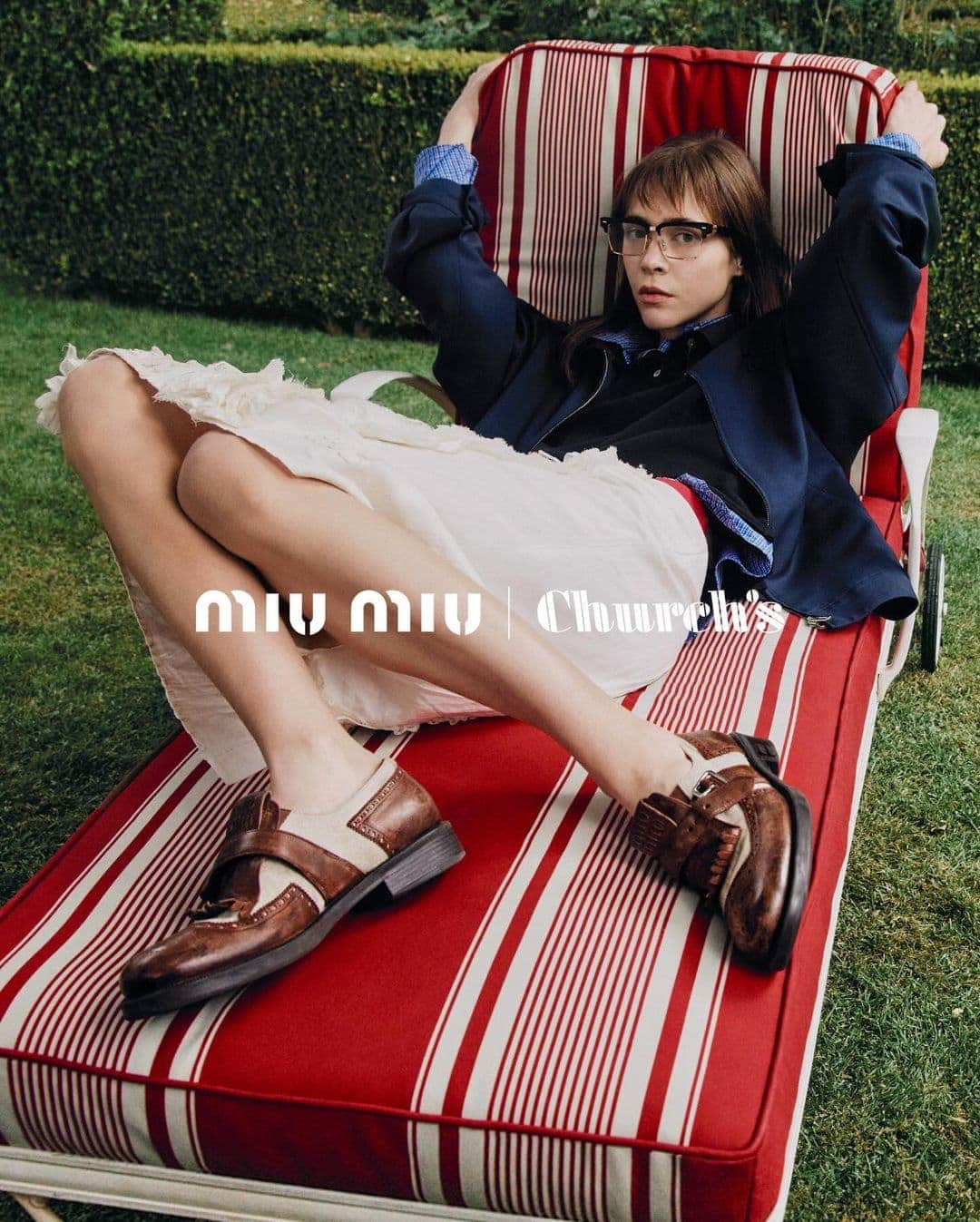 Young woman in glasses lounges on a red and white striped chaise, wearing a navy jacket, white skirt, and brown and white shoes outdoors.