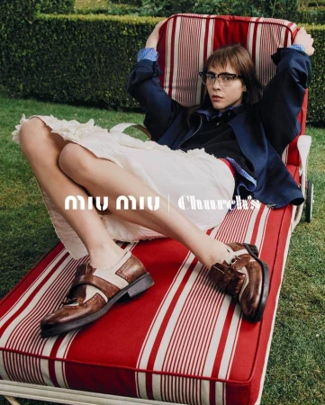 Young woman in glasses lounges on a red and white striped chaise, wearing a navy jacket, white skirt, and brown and white shoes outdoors.