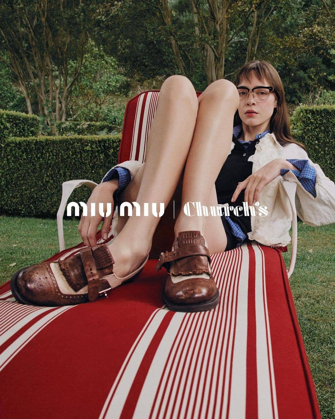 Fashion shot featuring woman on striped lounge chair, wearing glasses and Mary Jane shoes with brand logos in the foreground, trees behind.