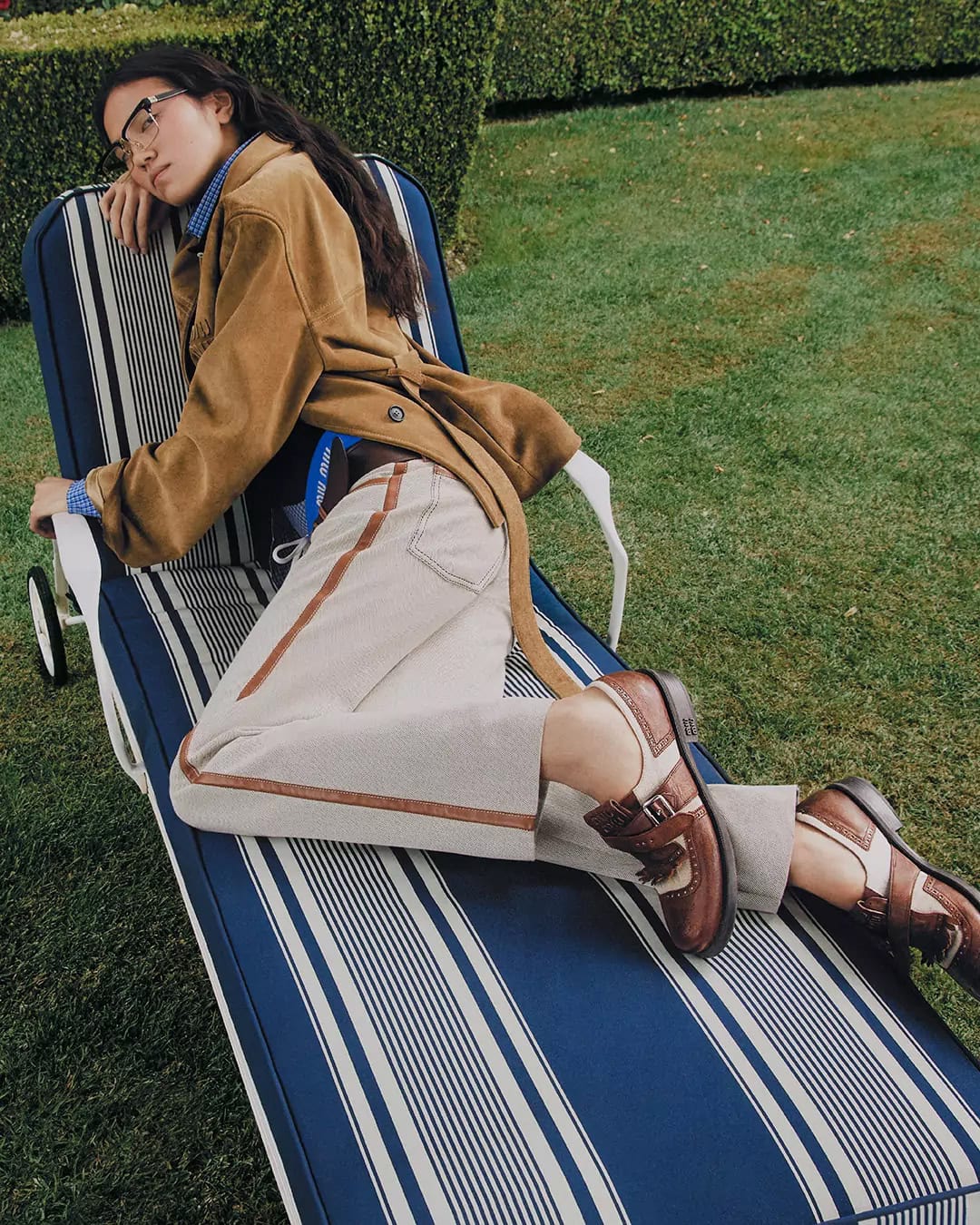 A young woman with glasses relaxes on a blue-and-white striped lounge chair in a grassy backyard, wearing a tan jacket and brown shoes.