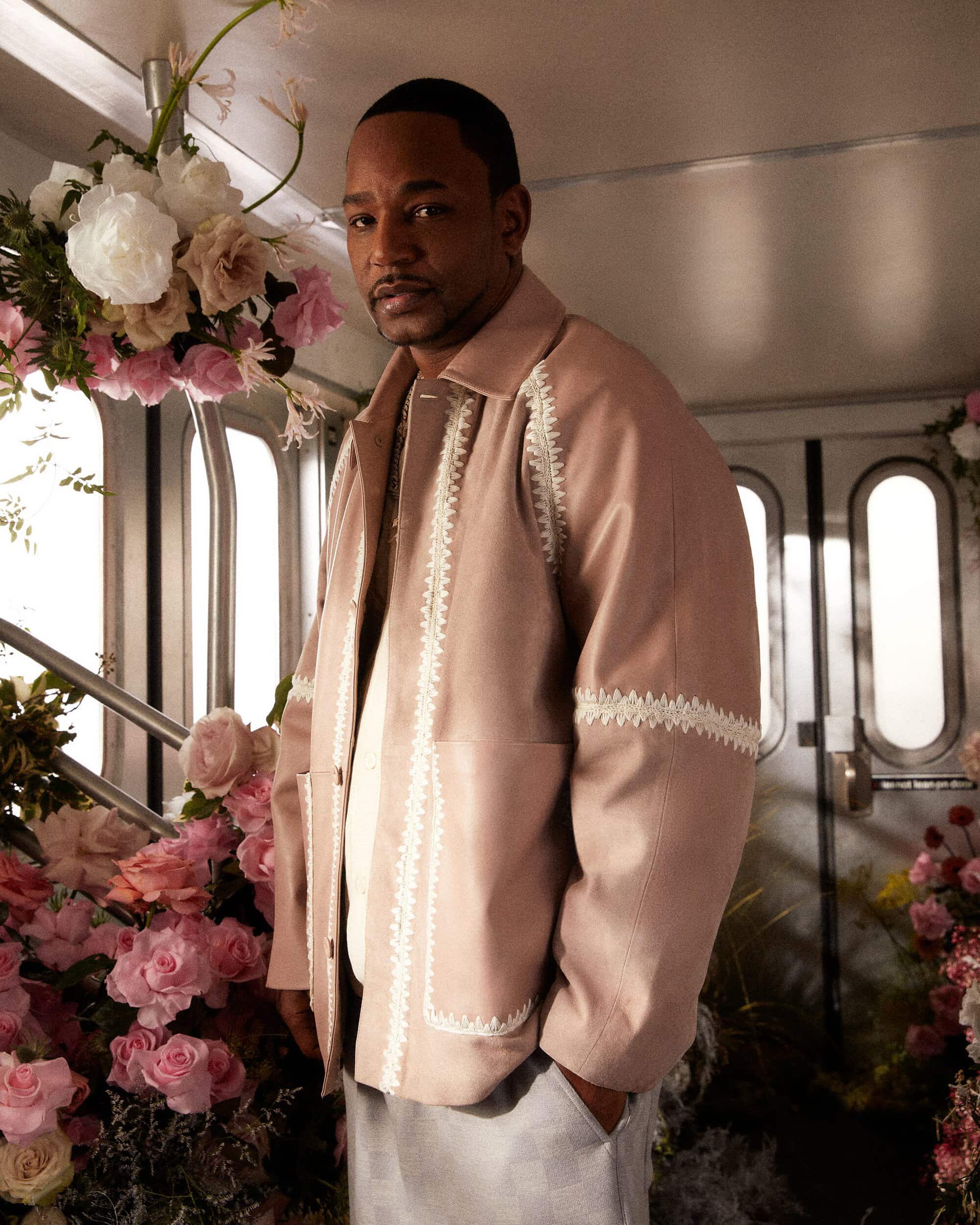Adult male poses in a light pink jacket with white trim, in front of a flower arrangement and paneled wall in an indoor setting.
